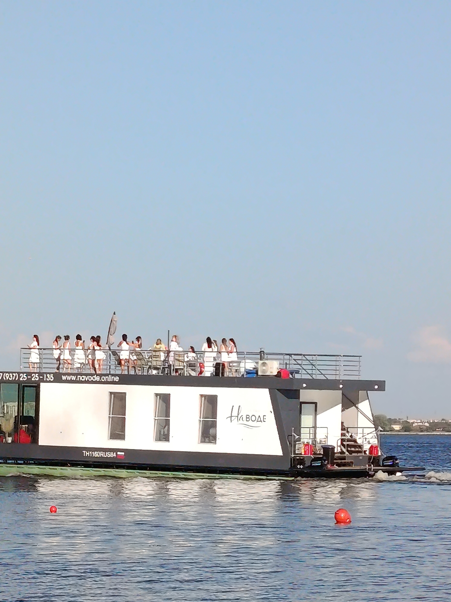 The image shows a wide, white houseboat with a black hull, featuring a deck crowded with people dressed in white. The houseboat has several windows on its side. Text on the side of the houseboat reads "НаВОДЕ" and "TH116DRUS04". On the deck, people are standing and sitting, some holding drinks. The houseboat is on a body of water, likely a river or lake, with a clear blue sky above. In the foreground, two orange buoys are visible in the water. The overall scene suggests a recreational outing or party on the water.