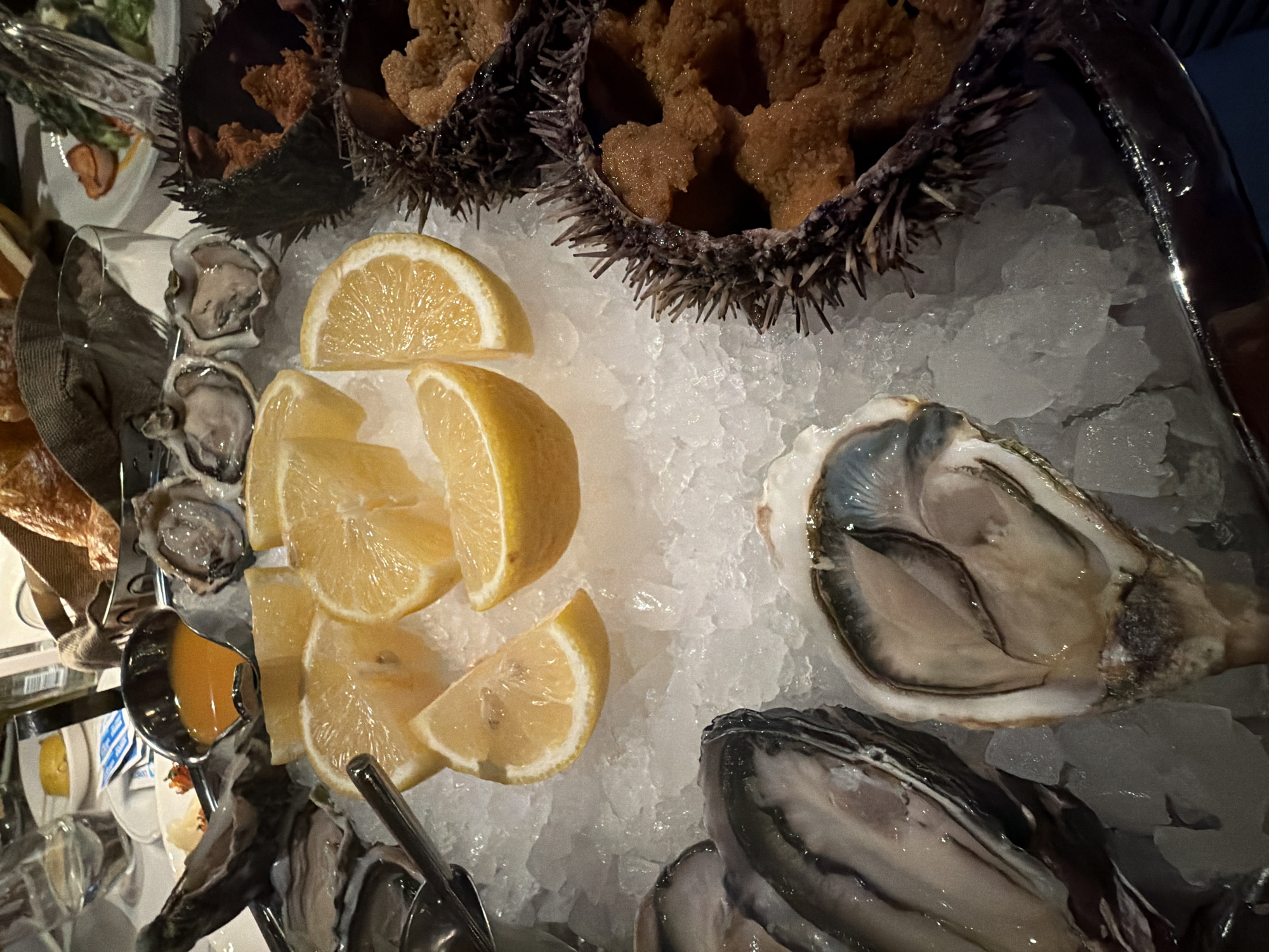 The image shows a platter of seafood on ice. At the top are two sea urchins, their shells filled with what appears to be roe. Adjacent to them, on the left, are several oysters. Slices of lemon are nestled between the oysters and the sea urchins. A small container with a sauce is visible on the left side. The platter is likely at a restaurant or bar in Moscow, Russia, as this type of seafood presentation is common in upscale establishments there.