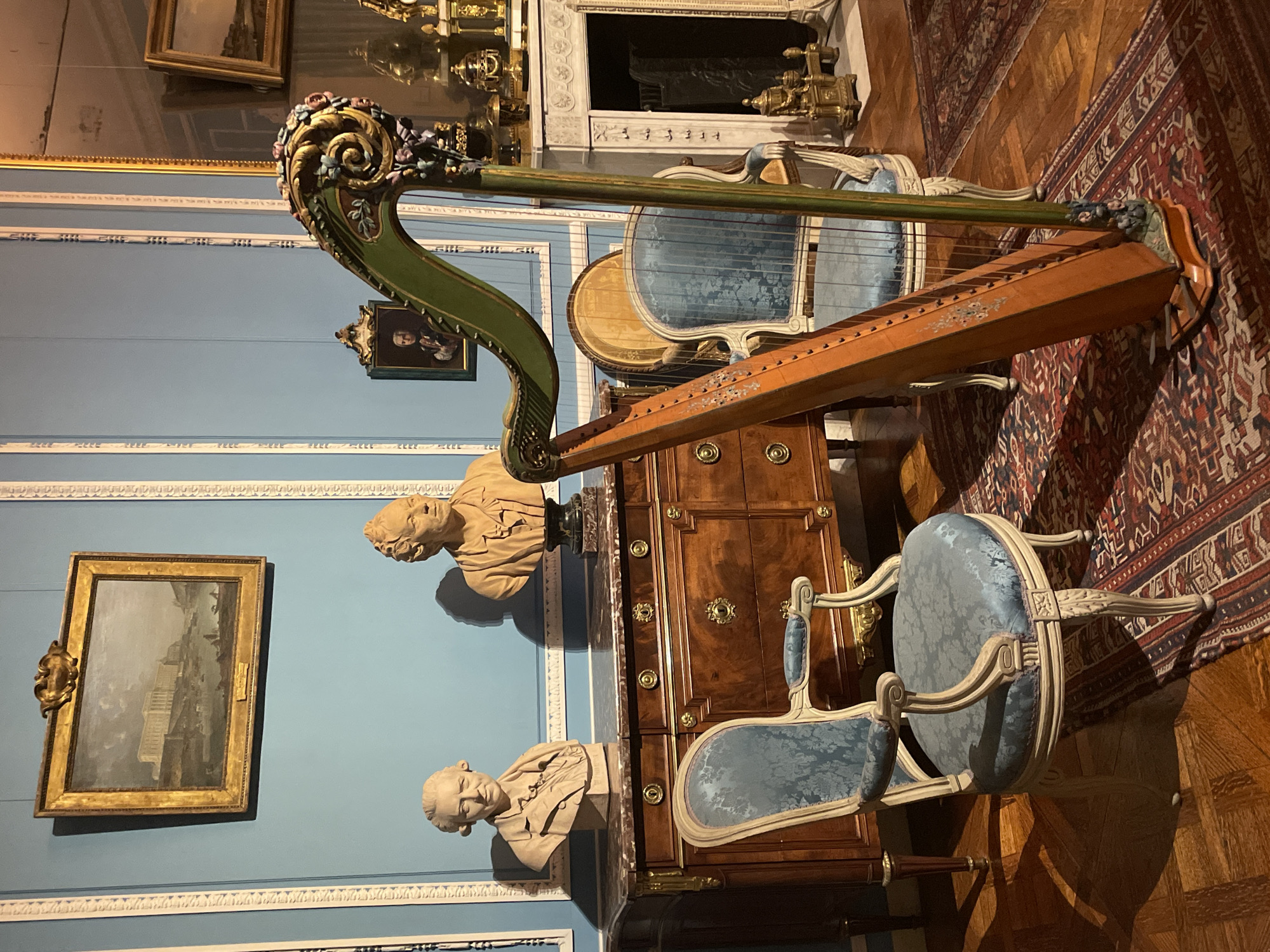 The image depicts an interior scene, likely a museum or historical room, showcasing a collection of antique furniture and decorative objects. A prominent feature is a wooden harp with a green, ornate pillar adorned with floral motifs. In front of the harp is a plush blue armchair with intricate carvings on its legs and arms. To the left of the harp and chair, a dark wooden chest of drawers with brass accents stands against a light blue wall with decorative molding. Two classical busts, one on the left and one on the right, are placed on top of the chest. Above the chest, a framed painting hangs on the wall. Further to the right, a fireplace with a mantelpiece displays various decorative items, and a large, ornate mirror reflects parts of the room. The floor is covered with a patterned rug laid over a wooden parquet floor. The overall impression is one of a well-preserved, elegant historical setting, possibly within a Parisian museum or a grand private residence. The scene is static, with no visible people or actions.