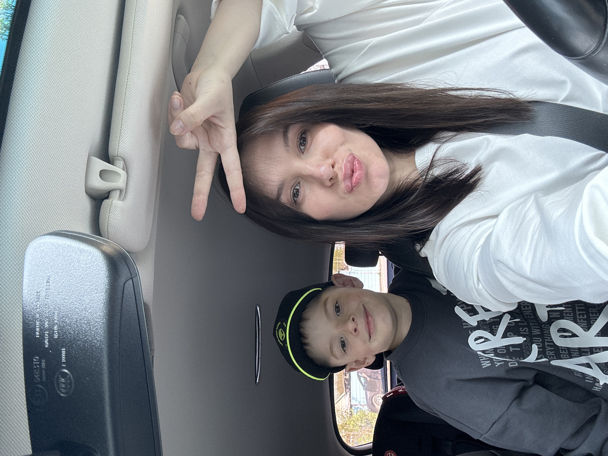 The image shows a woman and a child in the front seats of a car. The woman is on the left and is making a peace sign with her hand. She is wearing a white shirt and has brown hair. The child is on the right and is wearing a gray sweatshirt and a black cap with a lime green brim. The background is out of focus, but appears to be a street scene, possibly in the city of Chaikovsky, Russia. The rear view mirror is visible in the left foreground.