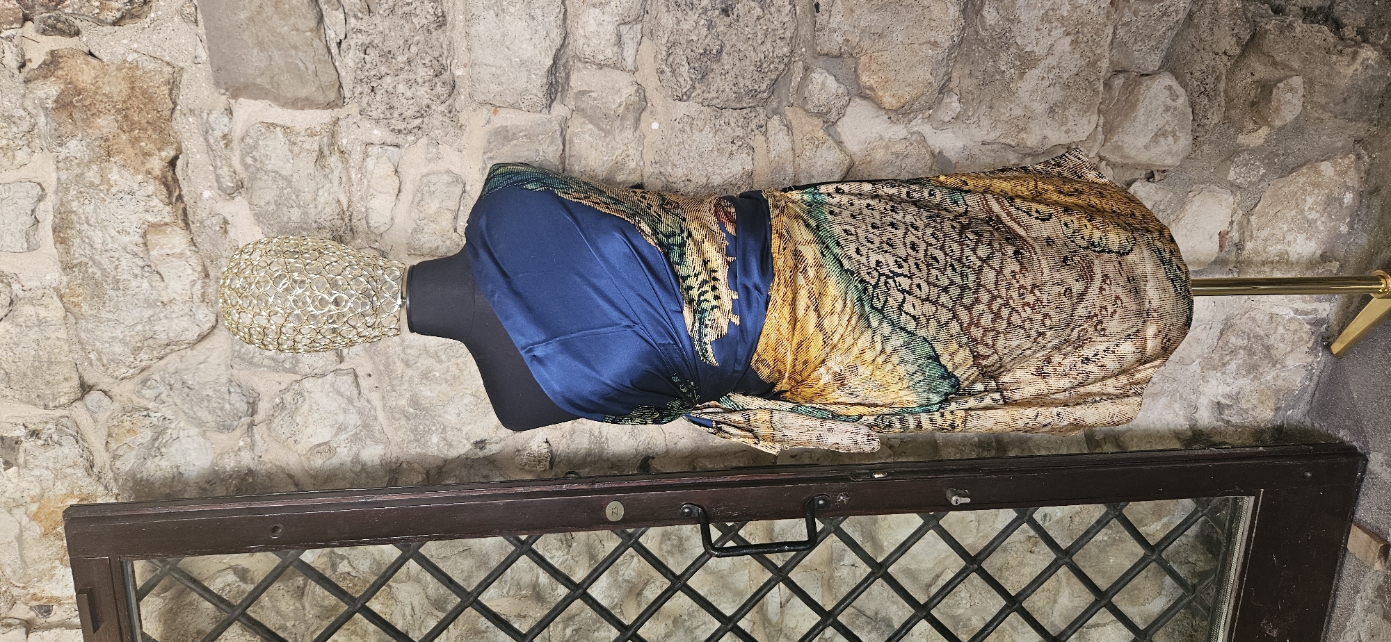 In Krakow, Poland, a mannequin is dressed in a draped garment featuring a blue satin fabric on the upper portion and a patterned fabric with animalistic and feather-like motifs in shades of gold, brown, green, and black on the lower portion. The mannequin is topped with a decorative gold cage-like headpiece. To the left of the mannequin stands a dark wooden door with a diamond-patterned glass pane and a metal handle. The backdrop consists of a rough stone wall. The scene is illuminated by artificial light, creating shadows.