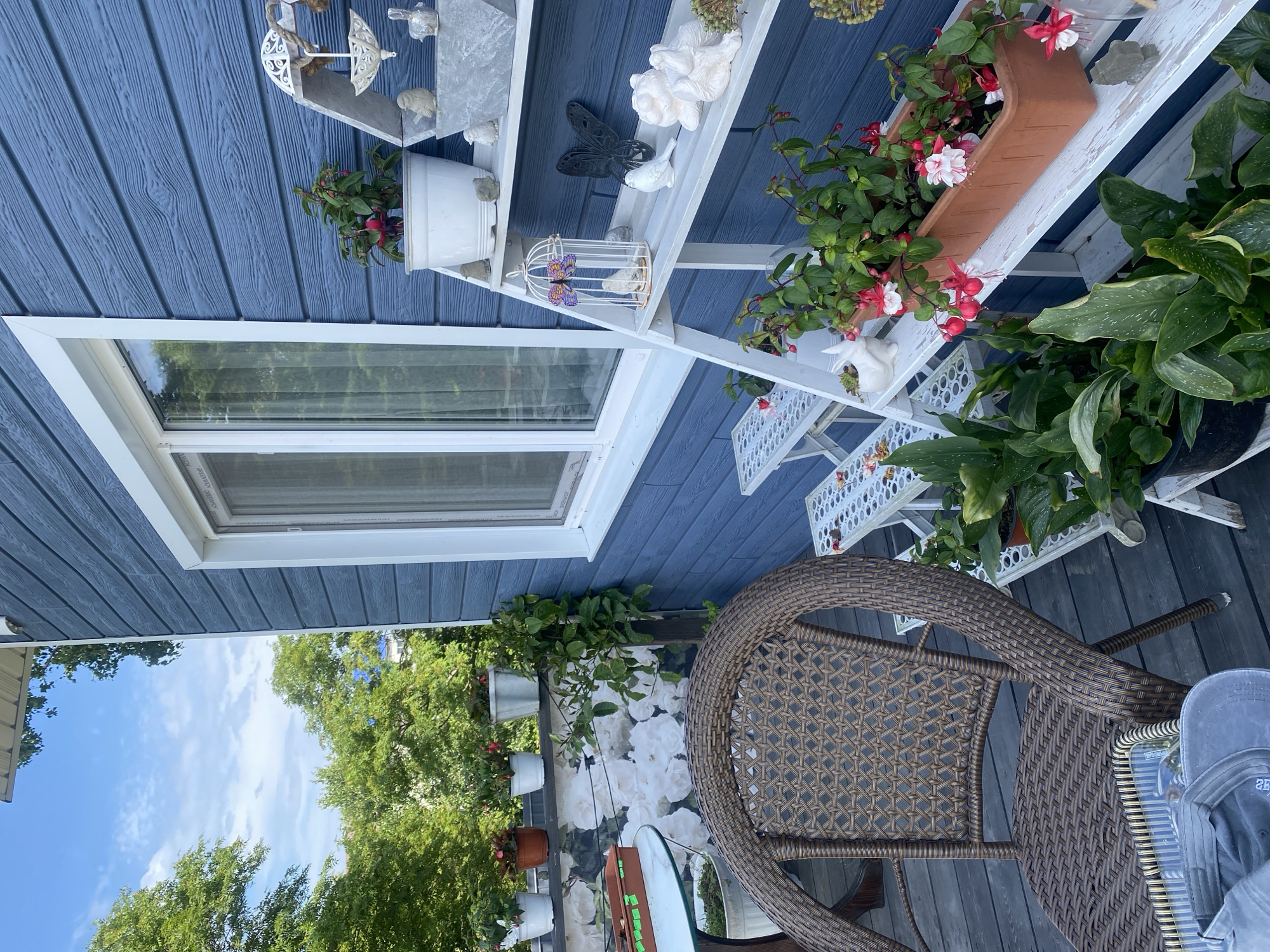 The image shows a close-up of a wooden deck in Tret’ya Pad’, Russia, featuring a blue wooden house with a white-trimmed window. To the right, a white tiered shelf unit displays various decorative items and potted plants, including fuschia. To the left, a wicker chair and a small table with a white floral-patterned backdrop are visible. The deck is furnished with a chair and a table, suggesting an outdoor seating area. Lush green trees and a partly cloudy sky form the background.