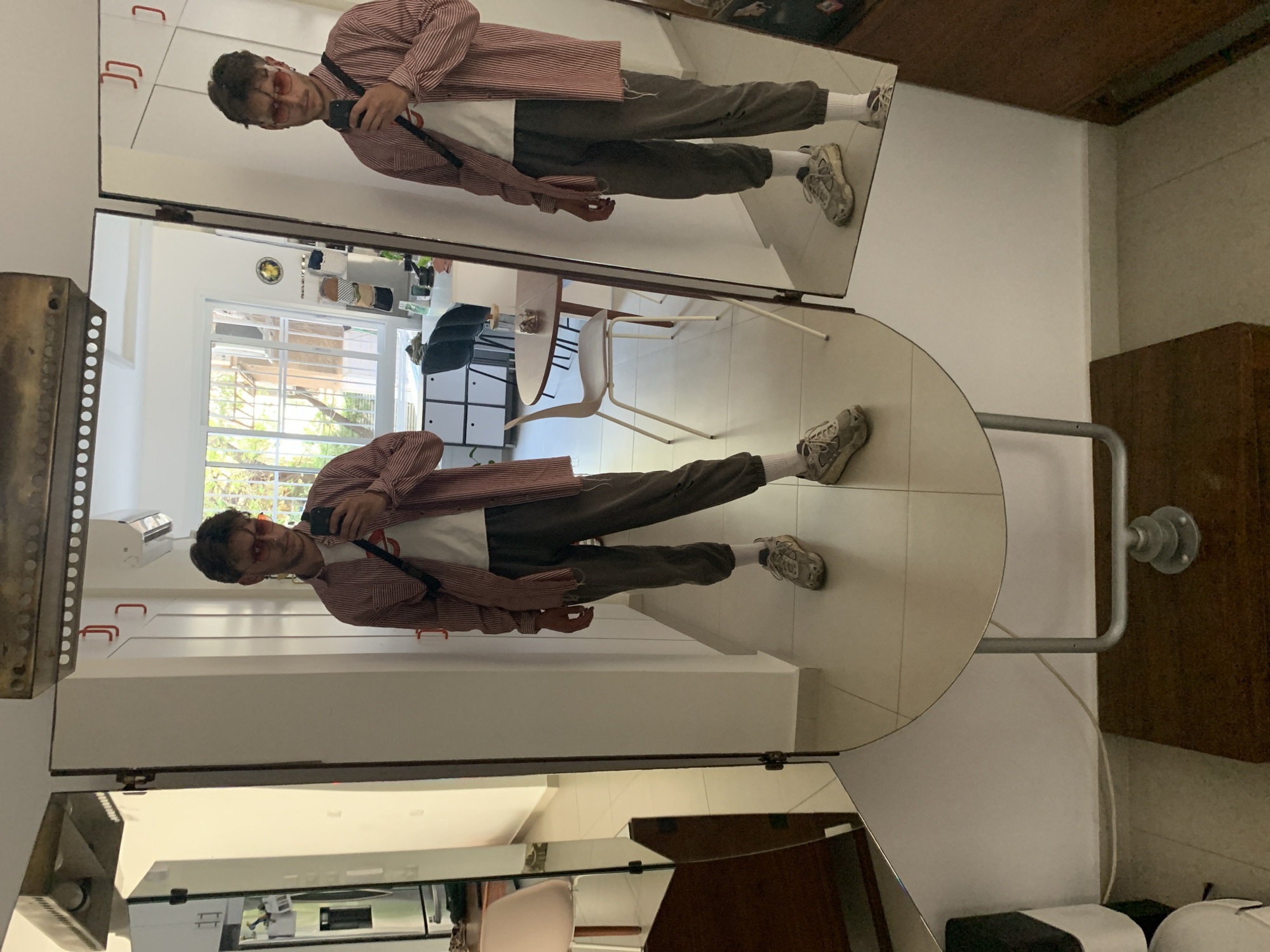 A vertical, medium shot depicts a man reflected in a multi-paneled mirror in a presumably indoor environment. The man, wearing a red and white striped shirt over a white t-shirt, grey pants, white socks, and grey sneakers, is holding a black smartphone, seemingly taking a photo of his reflection. He is also wearing orange-tinted sunglasses.
The multi-paneled mirror consists of three parts: a rectangular upper section, a circular middle section, and a rectangular lower section. The circular mirror's frame is visible, indicating it's a standalone piece.
In the background, reflections within the mirrors reveal elements of a room including a dining area with white chairs and a table, a window with visible greenery outside, and various furniture pieces. The flooring appears to be tiled in a light color. A white air conditioning unit is visible in the top left part of the mirror.
While the image's location cannot be definitively confirmed as Tel-Aviv, Israel based on the visible elements alone, there is no visual information that contradicts the provided location.