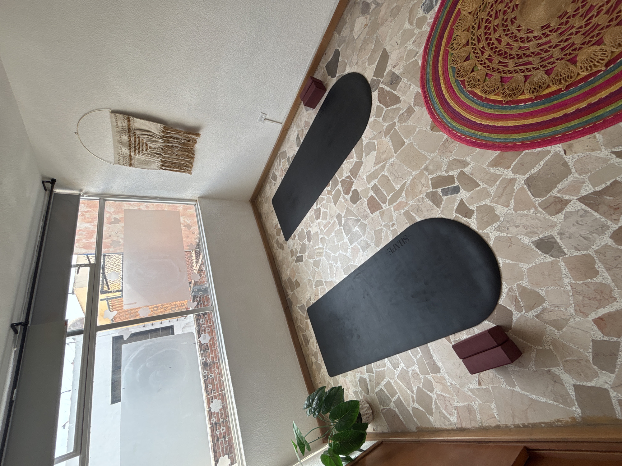 The image captures an indoor space, likely a studio or dedicated exercise room, set up for yoga or a similar activity. The scene is dominated by a distinctive floor made of irregularly shaped, light-colored stone or terrazzo tiles.
In the center, two black yoga mats are laid out, oriented vertically within the frame. The lower mat clearly displays the word "SHAPE" in white text. Adjacent to each mat are two burgundy-colored yoga blocks, neatly stacked, suggesting readiness for a session.
To the left, a large window extends from floor to ceiling, providing natural light. Portions of the window are frosted with decorative white cloudy patterns, offering privacy while allowing light to filter in. Through the clear sections of the window, glimpses of brickwork, metal railings, and other building structures can be seen outside, indicating an urban or semi-urban setting. A small green potted plant sits near the base of the window.
The upper wall features a woven, macrame-style wall hanging with fringes, attached to a semi-circular metal frame. In the upper right corner of the frame, a vibrant, circular woven rug or mat is partially visible, boasting concentric rings of red, pink, yellow, blue, and green, along with intricate golden patterns in its center. A white electrical outlet is visible on the upper wall.
The space appears clean and organized, with natural light suggesting it is daytime. No people are present, implying a pause before or after an activity. The combination of functional exercise equipment with decorative elements creates a serene and inviting atmosphere.