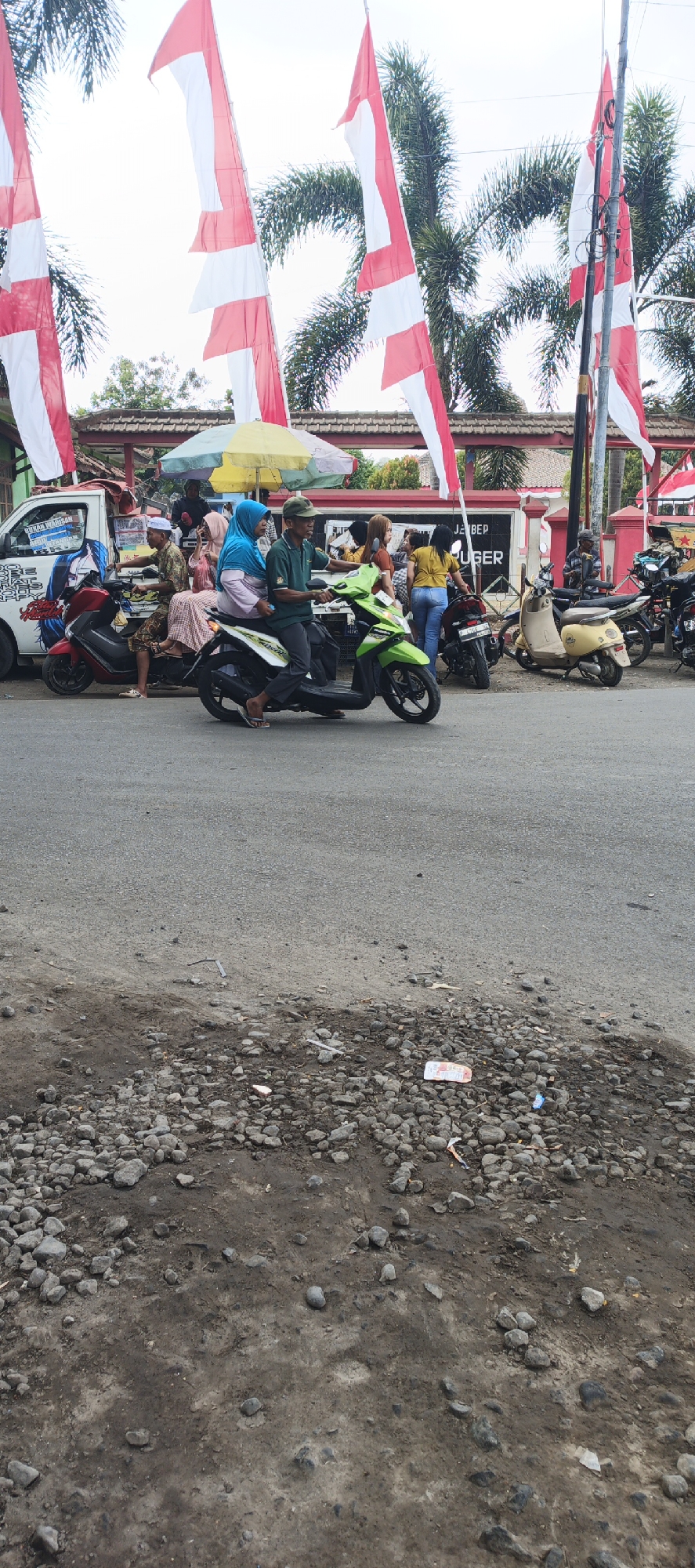 In Puger, Indonesia, a street scene unfolds with several people and vehicles. In the foreground, a dirt and gravel shoulder lines the asphalt road. On the road, a man in a green shirt and cap rides a green scooter, accompanied by a woman in a blue hijab. To their left, a red scooter is parked, and a man wearing a white cap sits on it. Further to the left, a white van with graphics is parked. Behind the scooters, several other individuals are standing or walking. Behind them, a building with a red facade and a tiled roof is visible. Several Indonesian flags, red and white, are prominently displayed on tall poles, interspersed with palm trees. To the right of the building, more motorcycles are parked, including a light yellow one. The scene appears to be a public area, possibly a market or a gathering point, with the flags suggesting a national celebration or event.