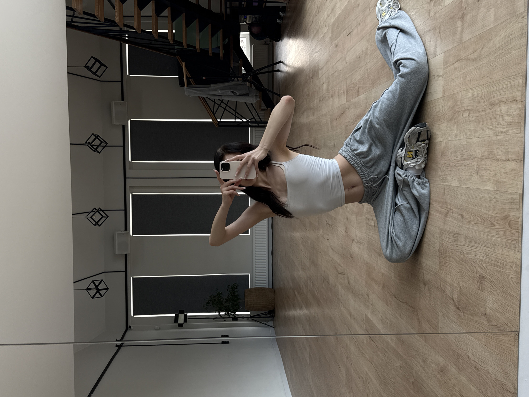 A person is taking a mirror selfie in a room with hardwood floors. The person is wearing a white tank top and grey sweatpants with athletic shoes. They are sitting with their legs extended. In the background, a staircase with a metal railing is visible. There are also windows with blinds, and some abstract geometric light fixtures hanging from the ceiling. A small plant in a basket is also in the background. The room appears to be a modern living space or studio. The lighting suggests it is daytime.