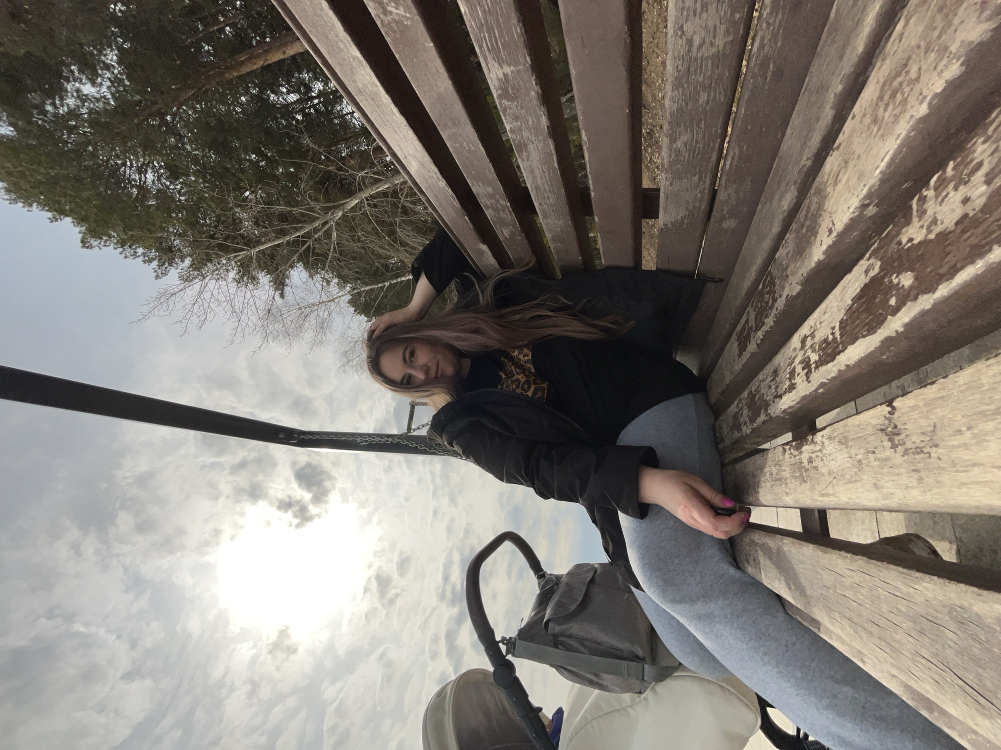 A woman is lounging on a weathered wooden park bench. She wears a black jacket over a printed top and grey leggings. She has long brown hair with lilac highlights. The sky is overcast with some sunlight breaking through. To the left is a stroller. In the background, there are trees, including a pine tree. The location may be Chaikovsky, Russia.