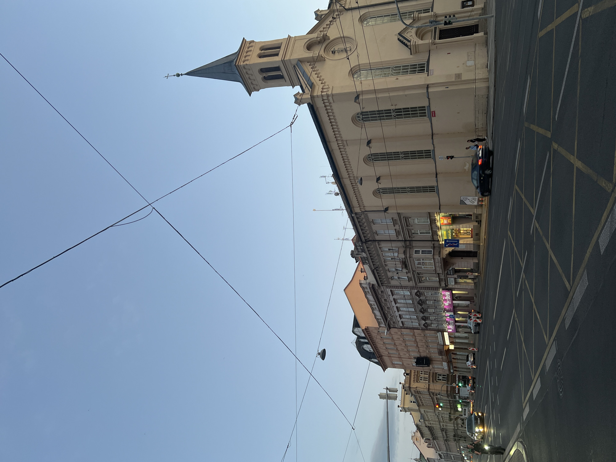 The image shows a wide street in the city of Pilsen, Czechia, during dusk. The sky is clear and blue, with a few clouds visible on the horizon. Several utility wires crisscross the sky. On the right side of the street is a large, light-colored building with a tall, pointed steeple, identified as the Evangelical Church of the Holy Spirit. The building has arched windows and decorative architectural details.
On the left side of the street, a row of multi-story buildings with various facades lines the road. Some of these buildings appear to be shops or businesses, with illuminated signs visible. The street itself is paved and features multiple lanes of traffic. Yellow and white road markings, including a box junction, are clearly visible. Several cars are present on the road, some in motion and some stopped. A few pedestrians can be seen on the sidewalk near the church. The overall impression is of a busy urban street scene as daylight fades.