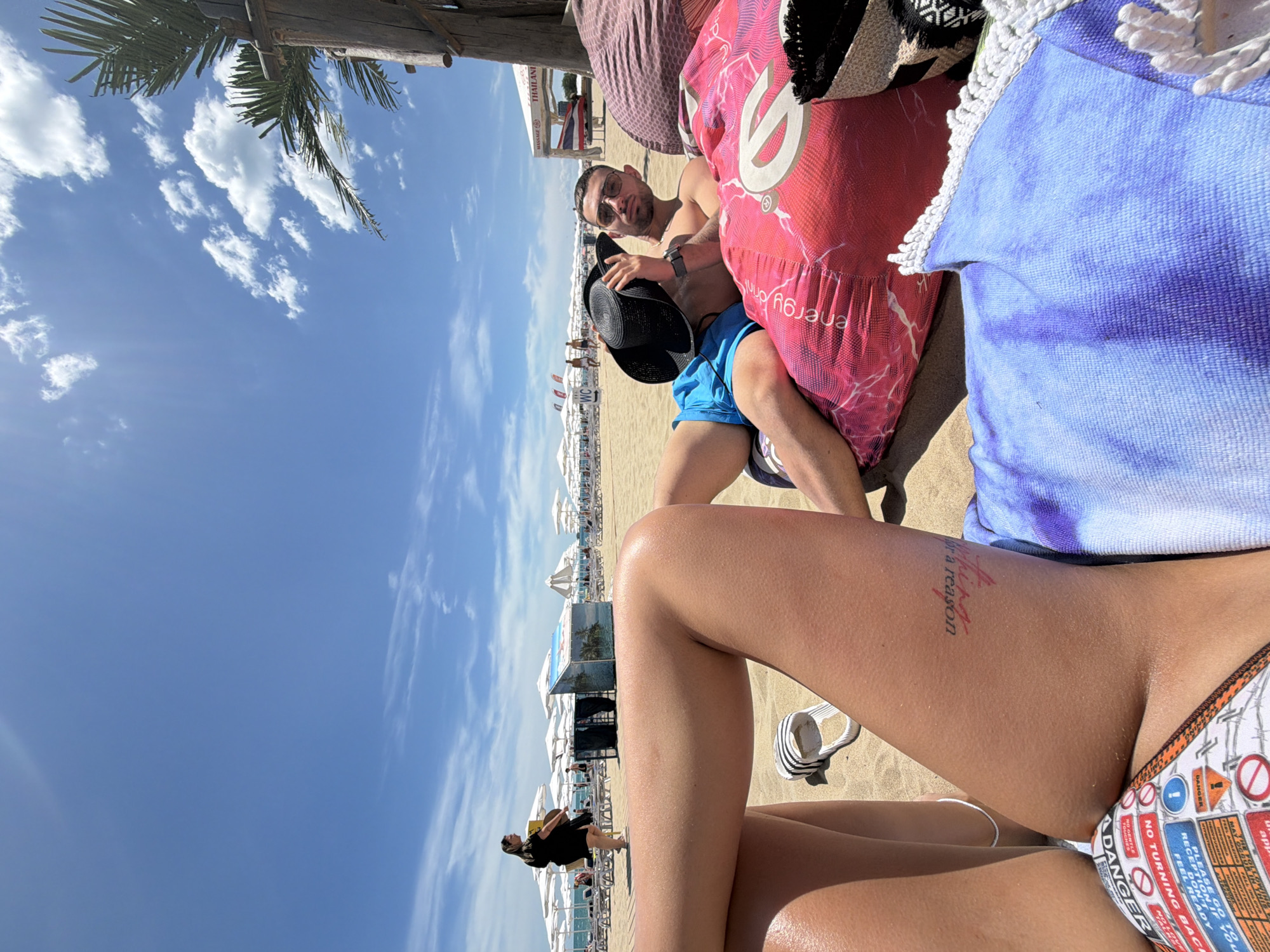 The image depicts a beach scene on a sunny day in Nesebar, Bulgaria. In the foreground, a person's legs are visible, wearing a bikini with a pattern of warning signs. To the right, a man wearing blue swim trunks and sunglasses is reclining on large beanbag chairs, holding a black hat over his face. He is partially obscured by the beanbag chairs, one of which is bright pink with a swirling pattern and text, and the other is a softer pink. Behind the couple, a sandy beach is lined with rows of white beach umbrellas and lounge chairs. In the distance, people are walking along the shore, and palm trees are visible, as well as a wooden structure with a sign that reads "MASSAGE THAILAND". The sky is blue with scattered white clouds.
