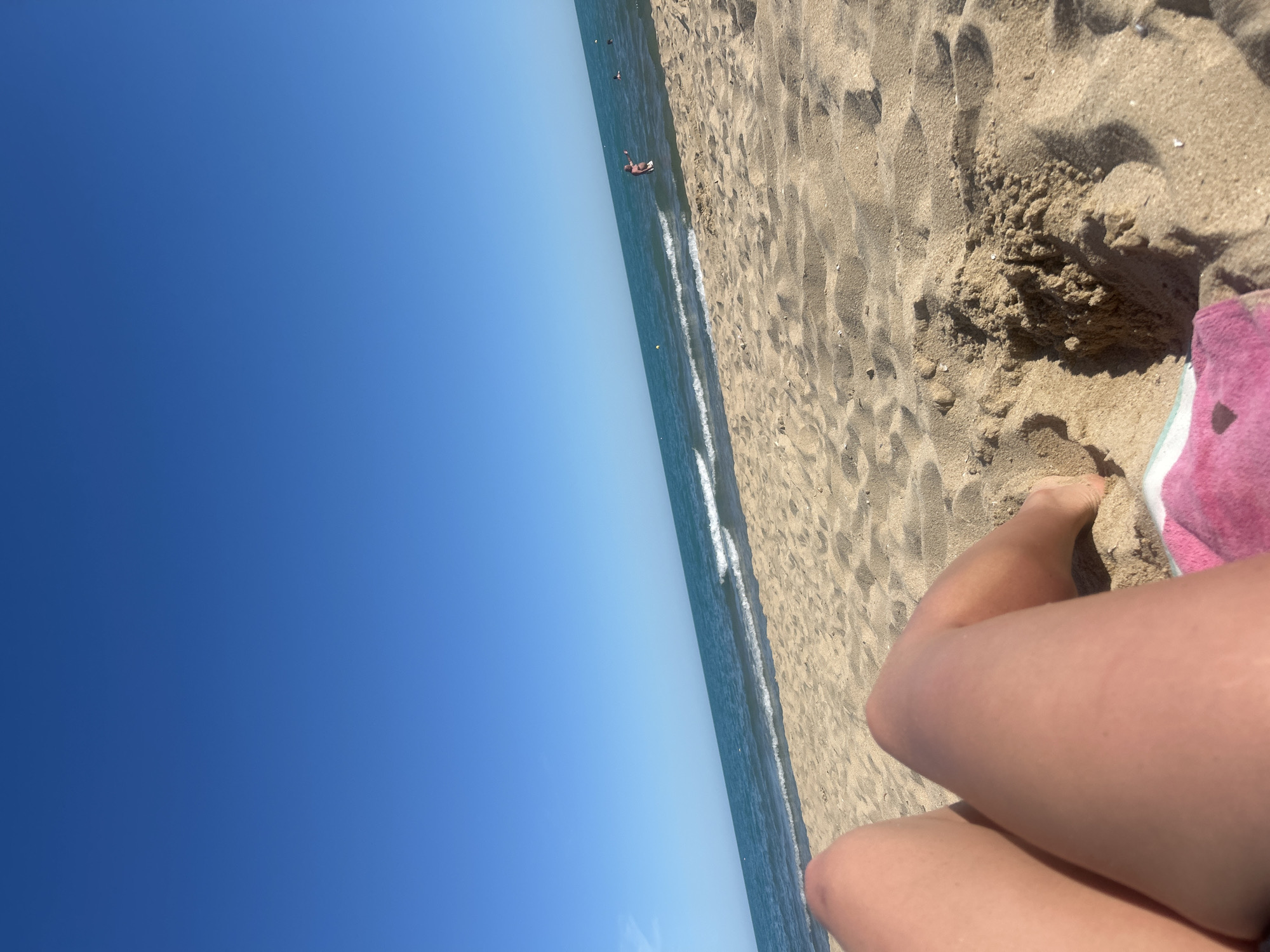 This media file captures a beach scene, likely in Varna, Bulgaria, under clear, sunny skies, suggesting a warm, pleasant day, possibly midday due to the bright sunlight.
The foreground is dominated by a person's legs and feet resting on the sandy beach, with a pink towel partially visible. The sand is textured with footprints and ripples, indicating recent activity. To the left of the sand, the turquoise sea meets the shore with small waves. Further out in the water, several distant figures can be seen, some swimming or wading. The sky is a vibrant, cloudless blue, stretching across the upper portion of the image and meeting the horizon of the sea. The overall atmosphere conveyed is one of relaxation and enjoyment of a typical summer beach day. There is no visible text in the image.