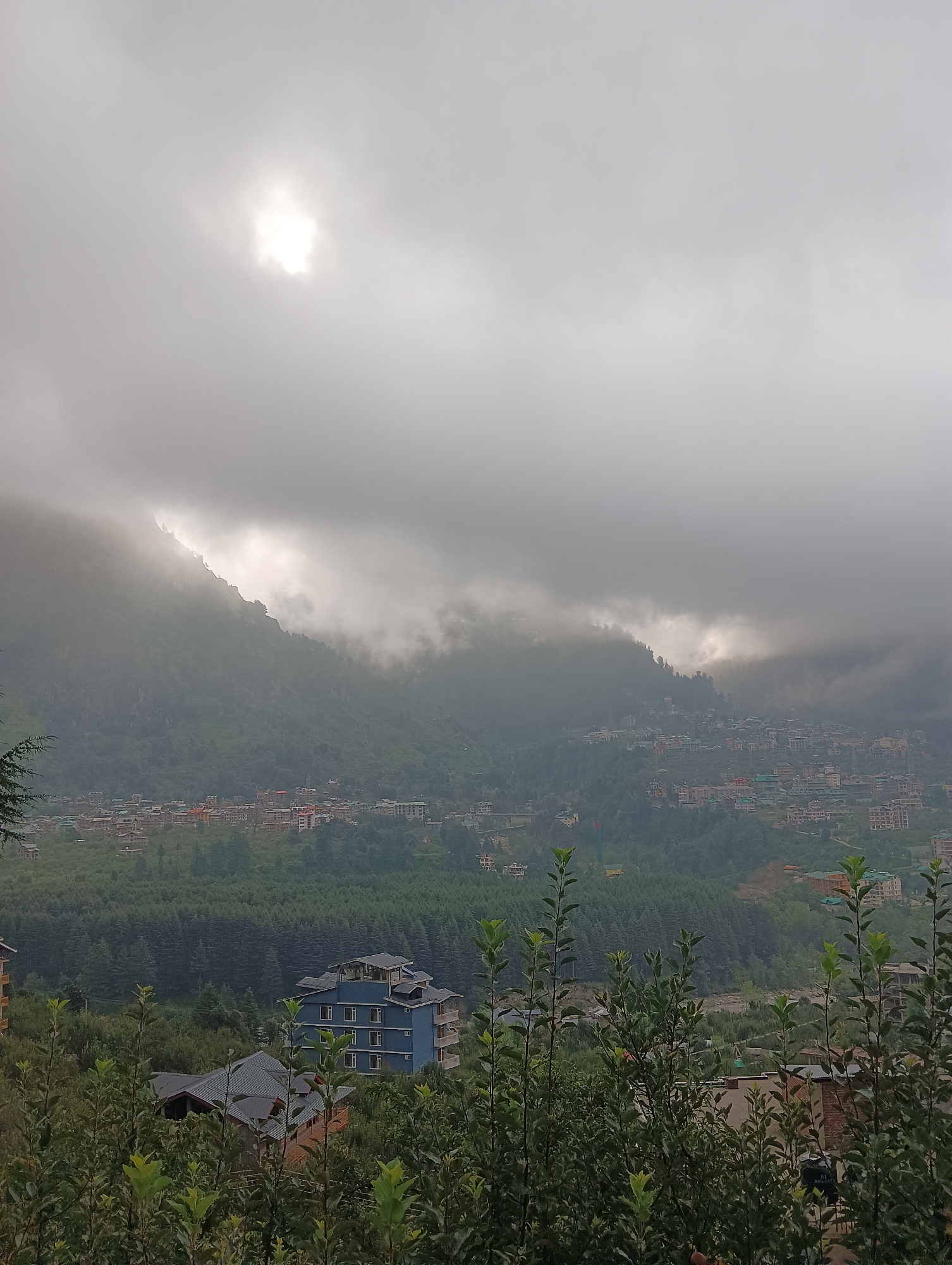 An overcast day is depicted in this panoramic view of a valley in Chhyal, India. The sun is a bright white orb breaking through a break in the thick, grey clouds, casting a diffused light onto a mountainside. In the middle ground, a dense forest of green trees covers the valley floor, with a small cluster of buildings visible in the distance. The buildings appear to be multi-story structures with red and white facades, nestled amongst the trees. The foreground is filled with lush green foliage, with the tops of trees and bushes creating a natural frame for the scene. The overall impression is one of a tranquil, rural landscape shrouded in mist and clouds.