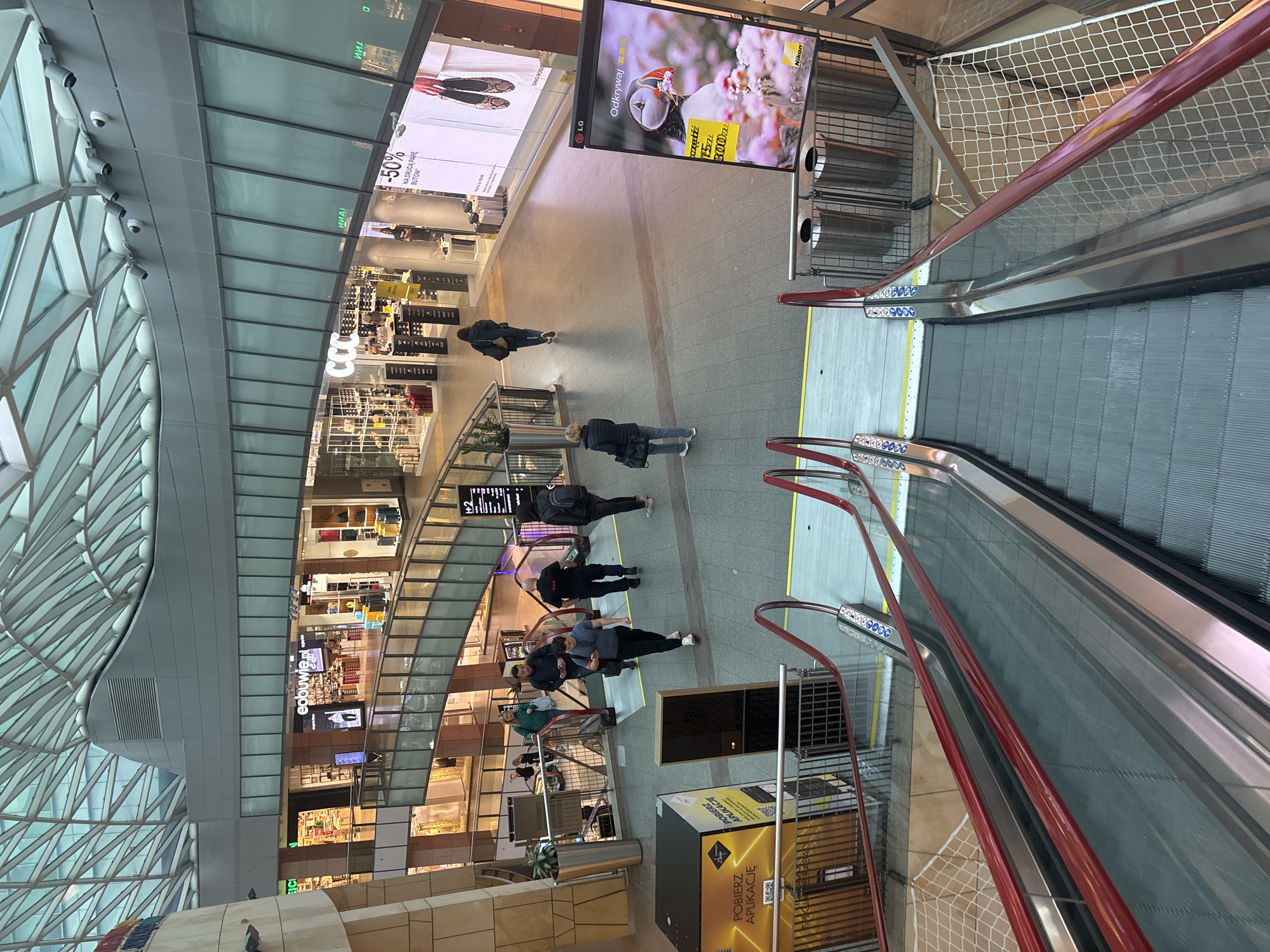 This image shows the interior of a shopping mall in Warsaw, Poland. The mall features a glass roof that allows natural light to illuminate the space. Several stores are visible on different levels, with brands like CCC and eobuwie.pl being identifiable. Multiple people are walking through the mall's central area. Two escalators with red handrails are prominent in the foreground, suggesting vertical movement between floors. Advertisements are displayed on large screens, with one advertising Nikon cameras. A sign promotes downloading an application. Overall, the scene depicts a typical day at a bustling shopping mall in Warsaw.