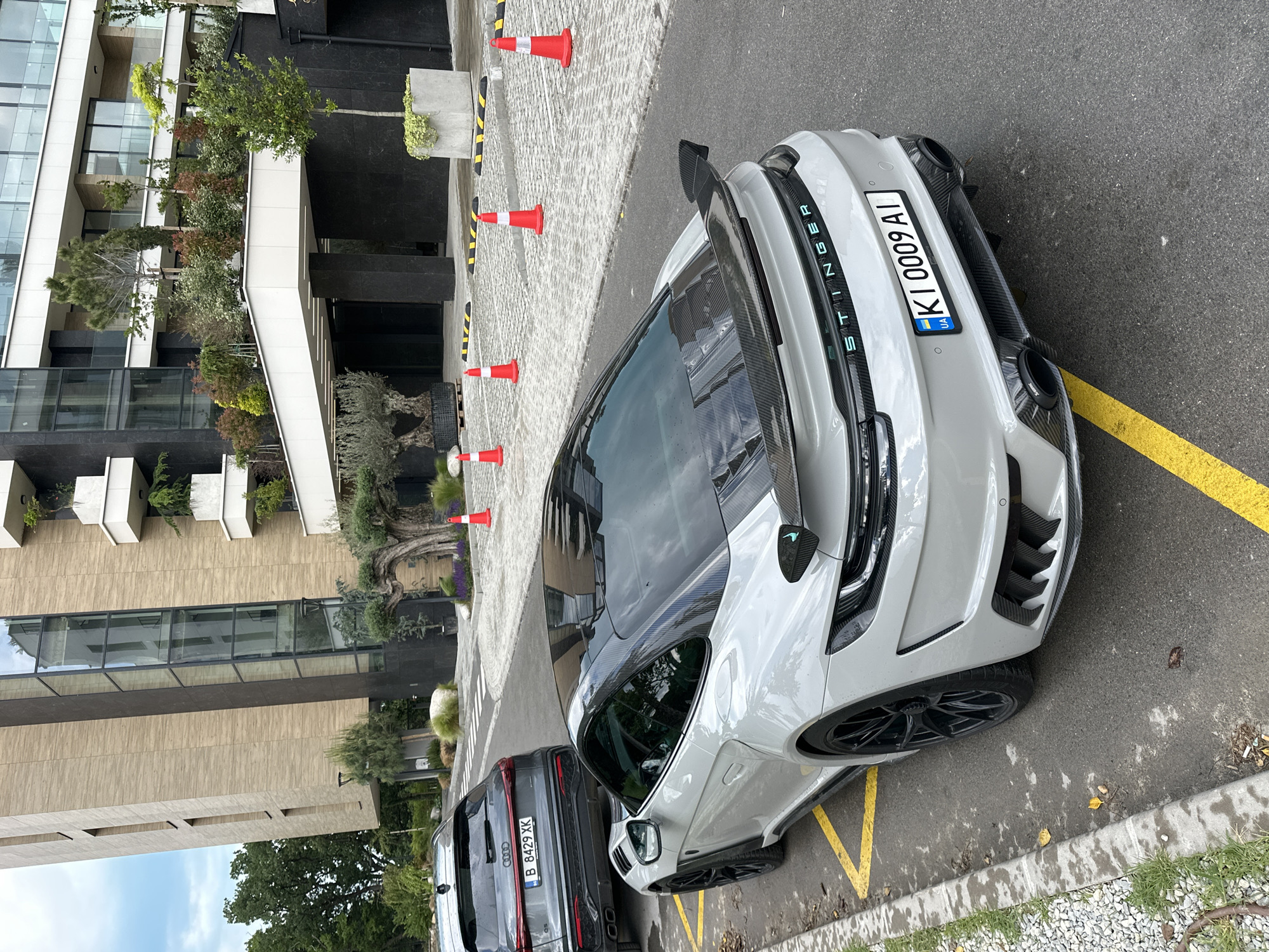 A grey Kia Stinger with license plate "KI 0009 AI" is parked near a modern building at an unknown location in Varna, Bulgaria. Orange traffic cones line the walkway leading to the building's entrance. A dark grey Audi SUV with license plate "B 8429 XK" is parked adjacent to the Stinger. The architectural style of the building is modern with visible plants on terraces. The environment includes a paved area, asphalt road, and some vegetation near the curb.