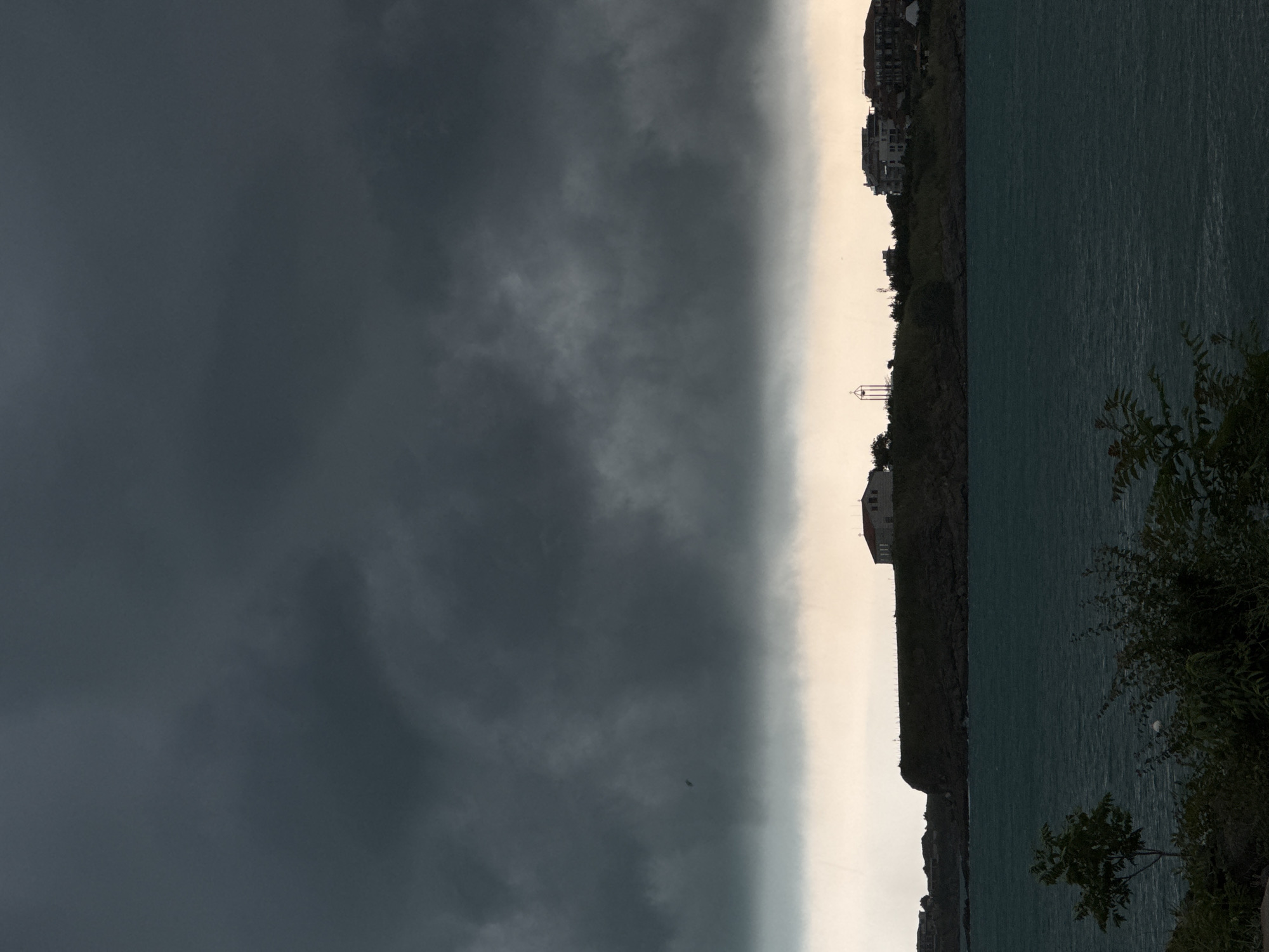 The image captures a dramatic coastal landscape under an impending storm, likely in Tsarevo, Bulgaria. The sky is dominated by a vast, dark, and brooding mass of storm clouds that occupies the upper-left portion of the frame. This dense cloud bank creates a stark, relatively horizontal boundary with a narrow band of much lighter, almost luminous pale gray sky stretching towards the right horizon.
Below this lighter sky, a serene body of water, presumably the Black Sea, fills the lower-right section of the image. The water appears deep teal to blue-green, with subtle ripples visible on its surface. Along the horizon, between the lighter sky and the water, lies a strip of land comprising dark, rugged cliffs or hills. Perched atop these elevated areas are several buildings, appearing to be houses or a small settlement, with one building featuring a distinctive white facade and a reddish roof. A small, white, tower-like structure, possibly a lighthouse or observation point, is also visible on the cliff edge. Sparse, dark green vegetation, including bushes and small trees, is present in the immediate foreground on the right.
A tiny, dark, indistinct object, possibly a bird or a distant drone, can be seen flying against the lighter sky portion. The stark contrast between the heavy, dark clouds and the lighter sky band suggests either late afternoon or early evening, or the moments just before a significant weather event. No people or visible text are present in the image.