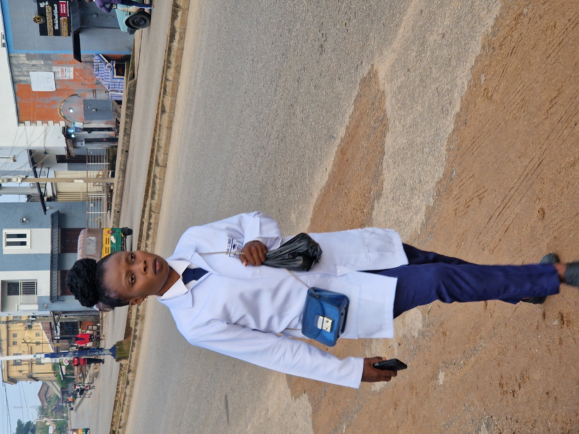 A medium shot captures a Black woman wearing a white lab coat, a dark blue tie, and dark blue trousers, walking on a dirt and paved road in Kabong, Nigeria. She has her hair styled in a high bun. She carries a small blue purse on her left side, a black plastic bag in her right hand, and a black smartphone in her left hand. The background shows a street scene with buildings, a road with vehicles including a rickshaw and a car, and a few other people. The lighting suggests it is daytime.