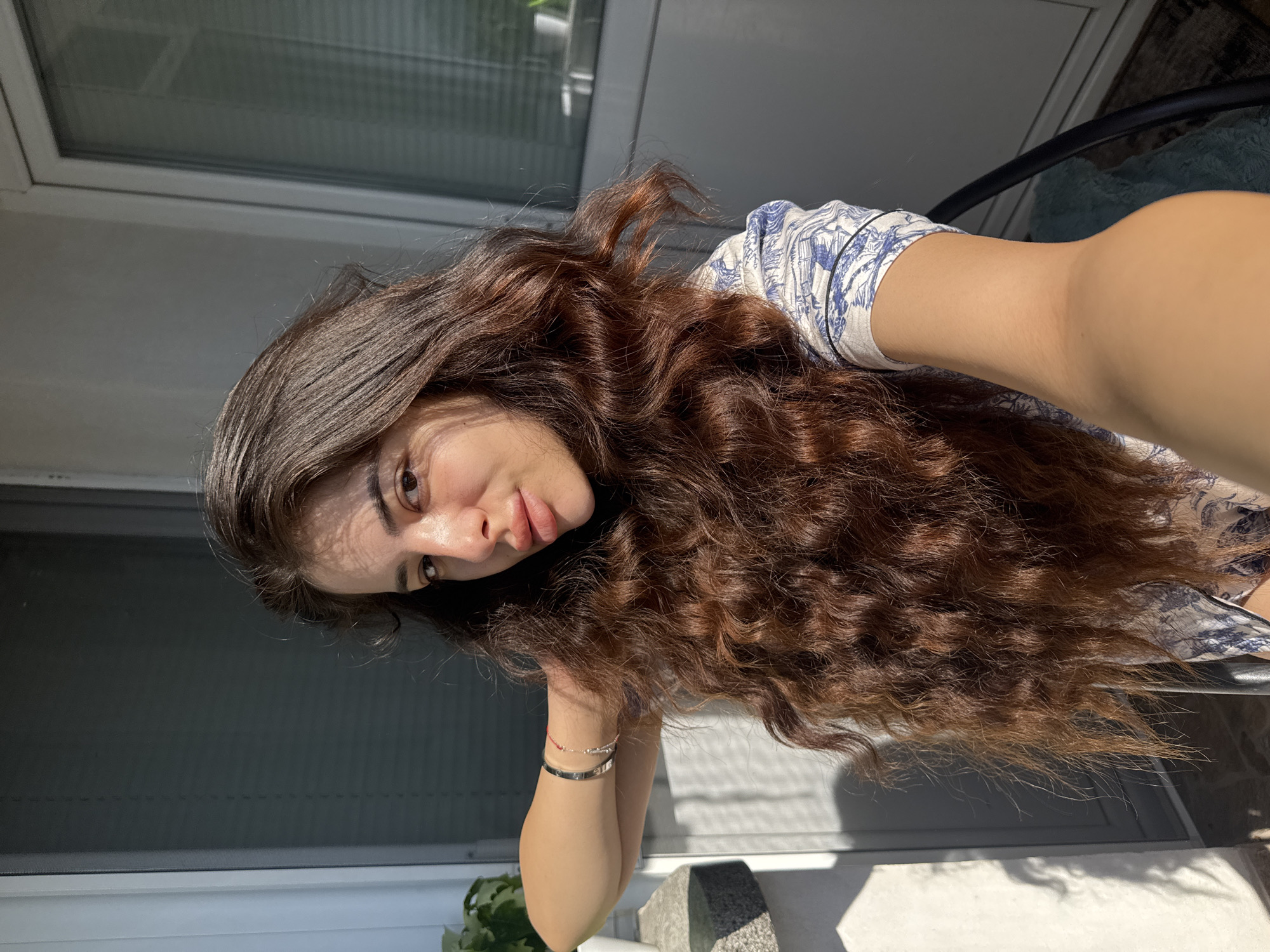 A young woman with long, wavy, dark brown hair is posing for a selfie on a balcony. She is wearing a blue and white patterned top. Her right arm is bent with her hand behind her neck, and her left arm is extended towards the camera, holding the phone. She is wearing a silver bracelet with a small red detail on her left wrist. The background shows a window with blinds and a plant on a ledge to the left, suggesting an urban setting, possibly in Plovdiv, Bulgaria, where this image was taken. Sunlight casts shadows across her face and the wall behind her.