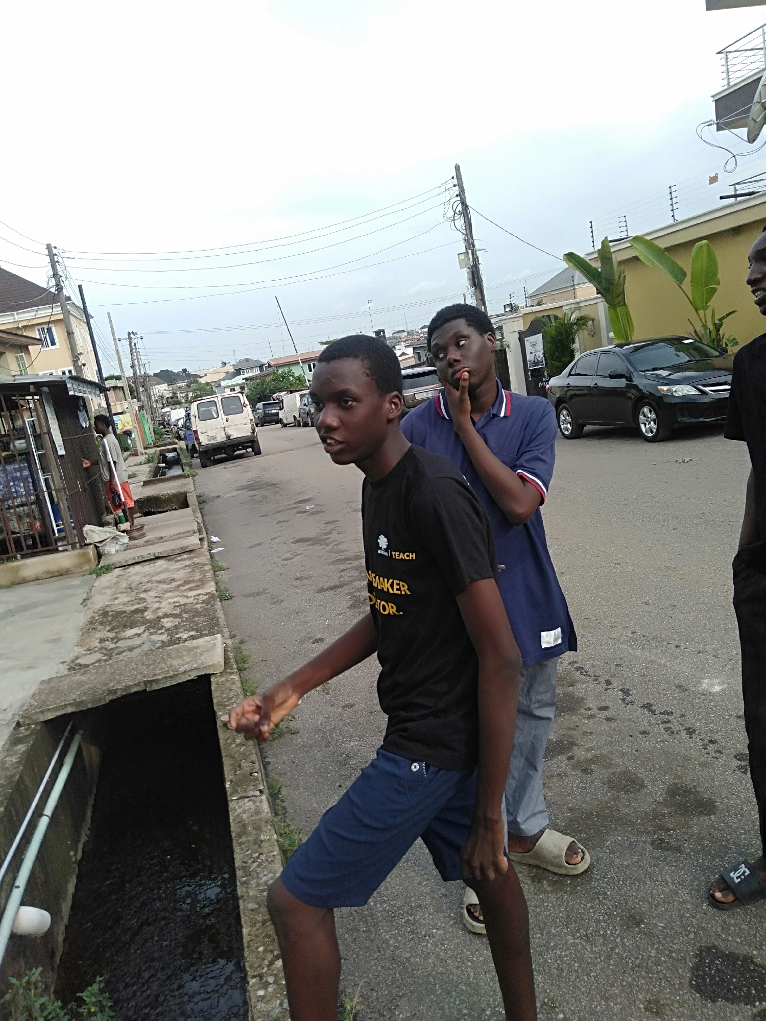 A street scene in an urban neighborhood features three young men. The central figure, clad in a black t-shirt and blue shorts, stands near a concrete drainage ditch, his hand resting on the edge. Behind him, another young man in a blue polo shirt appears pensive, his hand to his chin. The third young man is partially visible on the right edge, wearing black pants and sandals. In the background, the street includes houses, parked cars, utility poles with wires, and a white van. A small vendor stall is to the left of the frame.