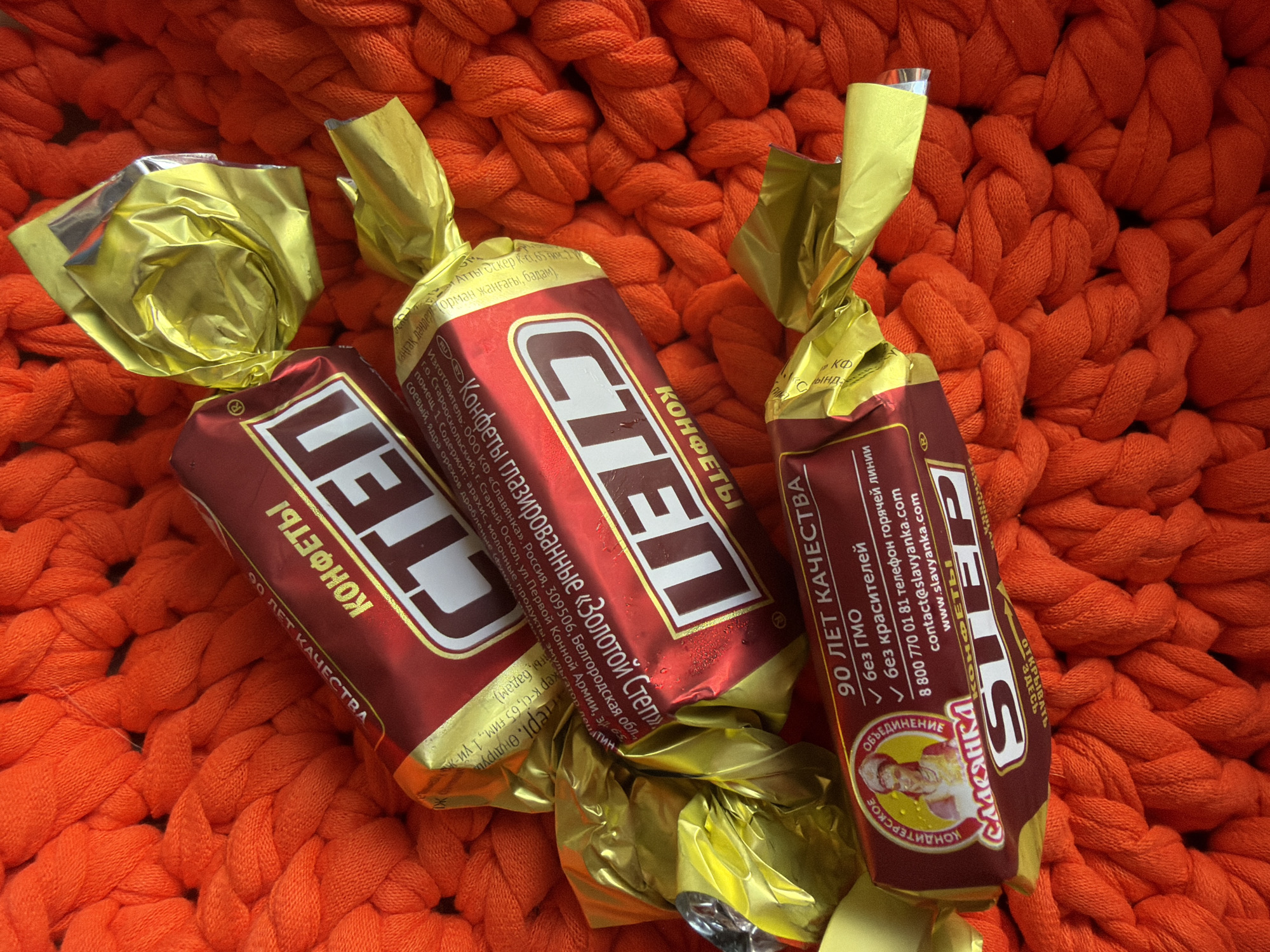 The image shows three individually wrapped candy bars, all with "STEP" prominently displayed on their wrappers in a bold, white font against a dark red background. The wrappers are a mix of dark red and gold foil. The candy bars are arranged on a textured, bright orange knitted surface.
Text on the wrappers is in Russian. One visible wrapper displays "КОНФЕТЫ" (candies) and "90 ЛЕТ КАЧЕСТВА" (90 years of quality). Another wrapper shows "КОНФЕТЫ глазированные" (glazed candies) and "ЗОЛОТОЙ СТЕП" (Golden Step). One wrapper features a circular logo with an image of a woman in traditional clothing, with the text "СЛАВЯНКА" (Slavyanka) above and "КОНДИТЕРСКОЕ ОБЪЕДИНЕНИЕ" (Confectionery Association) below. This wrapper also includes contact information: a phone number "8 800 770 01 81", an email "contact@slavyanka.com", and a website "www.slavyanka.com".
There is no indication of the city of Chaikovsky, Russia, in the image. The scene is a close-up, overhead view of the candy bars and the orange background.