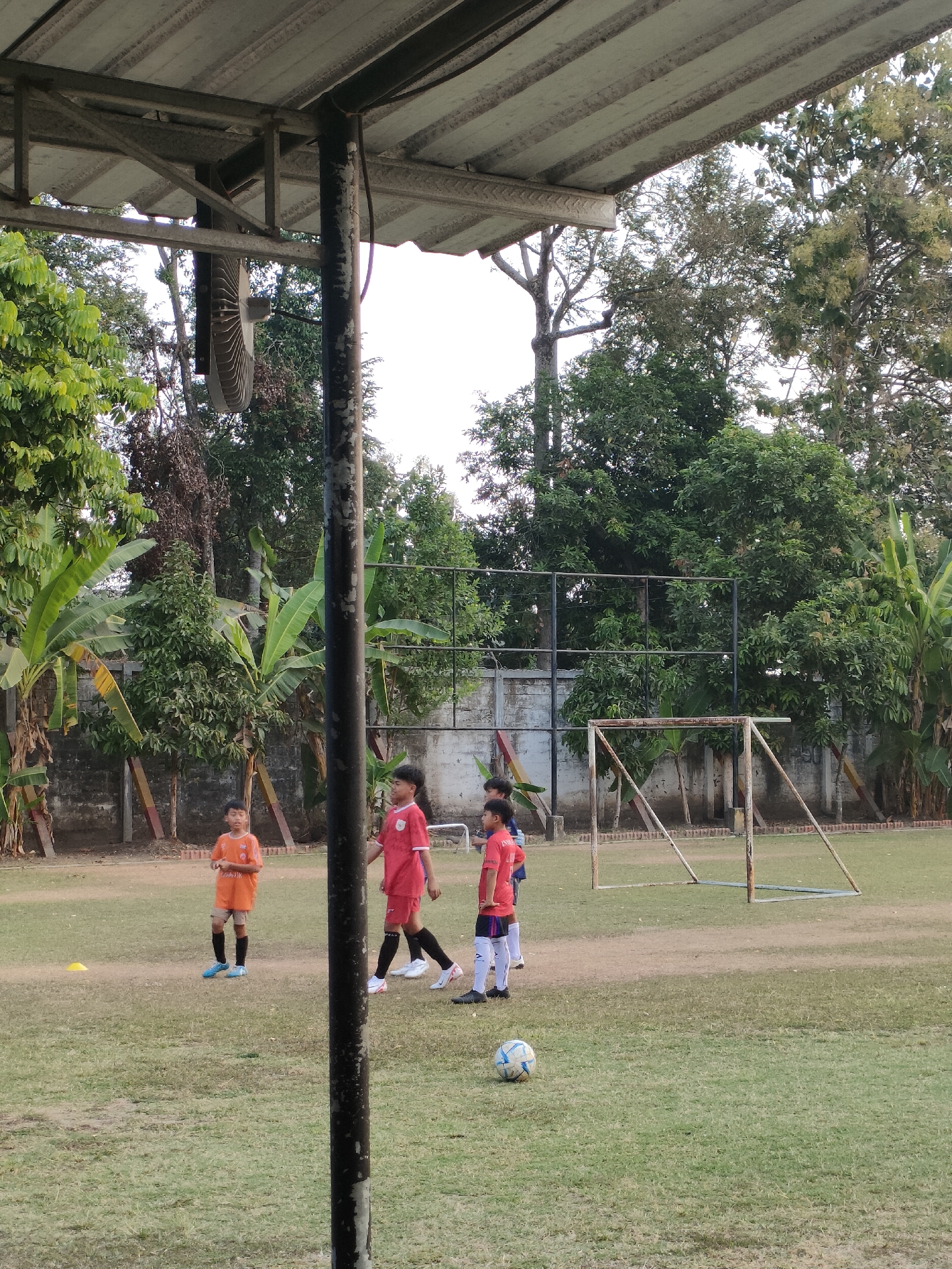 The image depicts an outdoor soccer practice for young children on a grassy field in Banguntapan, Indonesia. Four children, dressed in athletic wear, are present on the field. One child in an orange jersey stands near the left side of the frame, while three children in red jerseys are positioned towards the center and right. A soccer ball is visible on the grass in the foreground. In the background, a soccer goal and a fence are present, surrounded by trees and vegetation. The scene is partially obscured by a large, dark, vertical pole in the foreground. A ceiling structure with a fan is visible at the top of the image. The lighting suggests it is late afternoon or early morning.