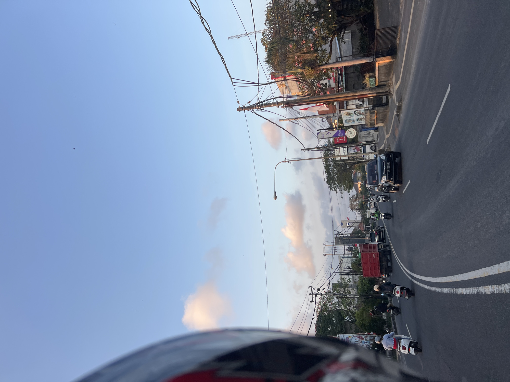 This is a POV shot from a motorcycle helmet looking out at a busy street in Denpasar, Indonesia. The road is paved with asphalt and marked with white lines. Several motorcycles and a red truck are visible on the road, along with a black SUV. On the right side of the road, there are buildings with signs, trees, and power lines. The sky is blue with a few scattered clouds, and the lighting suggests it is either early morning or late afternoon.