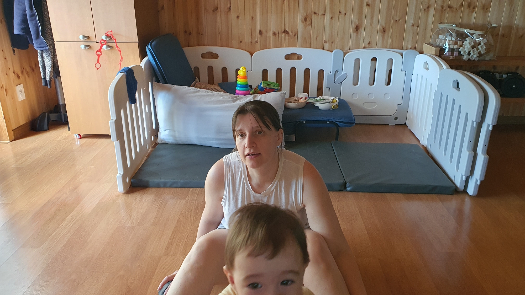 The image shows a woman sitting on a wooden floor, with a baby in front of her. The woman is wearing a white tank top. Behind her is a play area enclosed by white plastic fencing, with gray mats on the floor. Inside the play area are items such as a white pillow, a stack of colorful rings, and a small table with bowls and spoons. In the background are wooden paneled walls and a light-colored cabinet. The image was taken in Dunnae-myeon, South Korea.