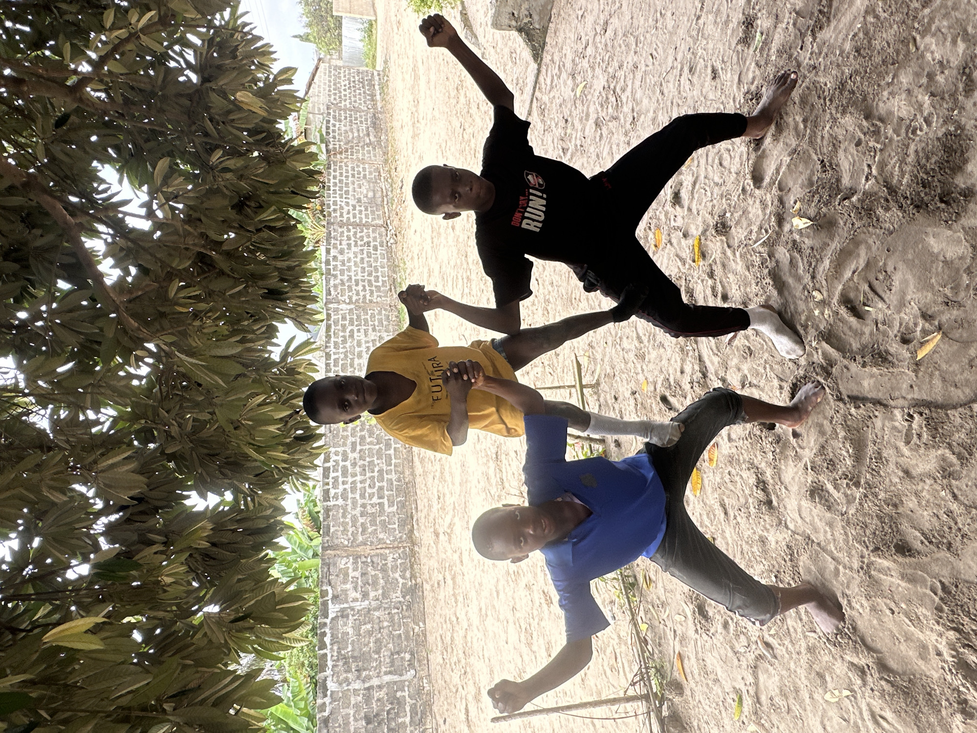 Three young boys are posing for a photograph in a sandy area beneath a tree. The boy on the left wears a blue polo shirt and dark pants. The boy in the middle wears a yellow t-shirt. The boy on the right wears a black t-shirt with white lettering that reads "DON'T SAY RUN!" and black pants. They are all making assertive poses with their fists raised. In the background, a low, light-colored brick wall borders the area, with some vegetation visible beyond it. The ground is covered in sand with a few fallen leaves. The scene appears to be an outdoor, informal setting, likely in a residential area of Badagry, Nigeria.
