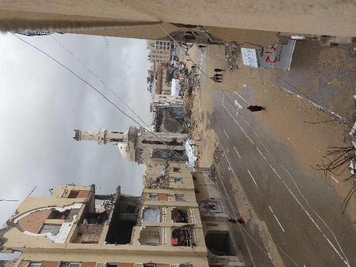 The image depicts an urban street scene of extensive structural damage in Khan Yunis, Palestinian Territory, under an overcast sky. On the left, a multi-story building is severely compromised, featuring large sections of its facade missing, exposing internal rooms, shattered windows, and debris on balconies. A mosque with a tall, slender minaret and a prominent dome stands in the mid-ground, showing considerable structural damage, including visible punctures and partial collapses, with its base surrounded by extensive piles of rubble and sand. A paved asphalt road, partially damp and coated with sand and dirt, traverses the center, marked with white double solid lines and a white right-turn arrow. Five individuals are discernible: one person in dark attire walks alone on the road, a second person in dark attire walks on the left sidewalk adjacent to the damaged building, a group of three to four individuals walks further down the street in the background, and an additional solitary figure is visible in the distant background. Electrical and communication wires are suspended across the upper and middle sections of the frame. A damaged building wall on the right partially frames the scene, displaying a white sign with red Arabic text.