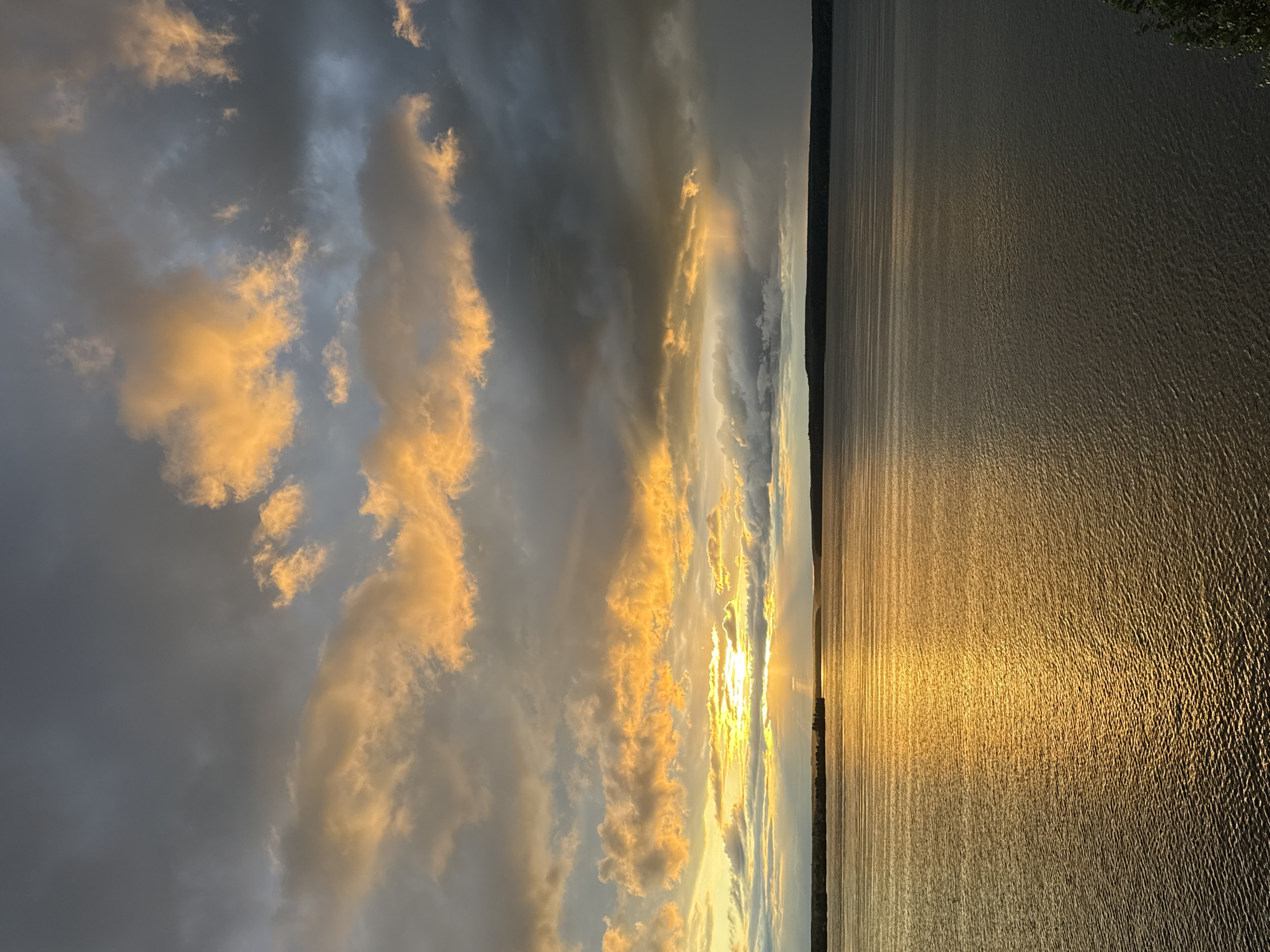 The image captures a sunset over the Kama River near Votkinsk, Russia. The sky is filled with dramatic clouds, some tinged with vibrant orange and yellow from the setting sun, while others are dark and ominous. Rays of sunlight pierce through the clouds, illuminating a path across the rippling water. The horizon line is marked by a dark, tree-covered shoreline. The overall atmosphere is one of natural beauty and the powerful forces of weather. There are no people or man-made structures visible in the image, focusing solely on the landscape.