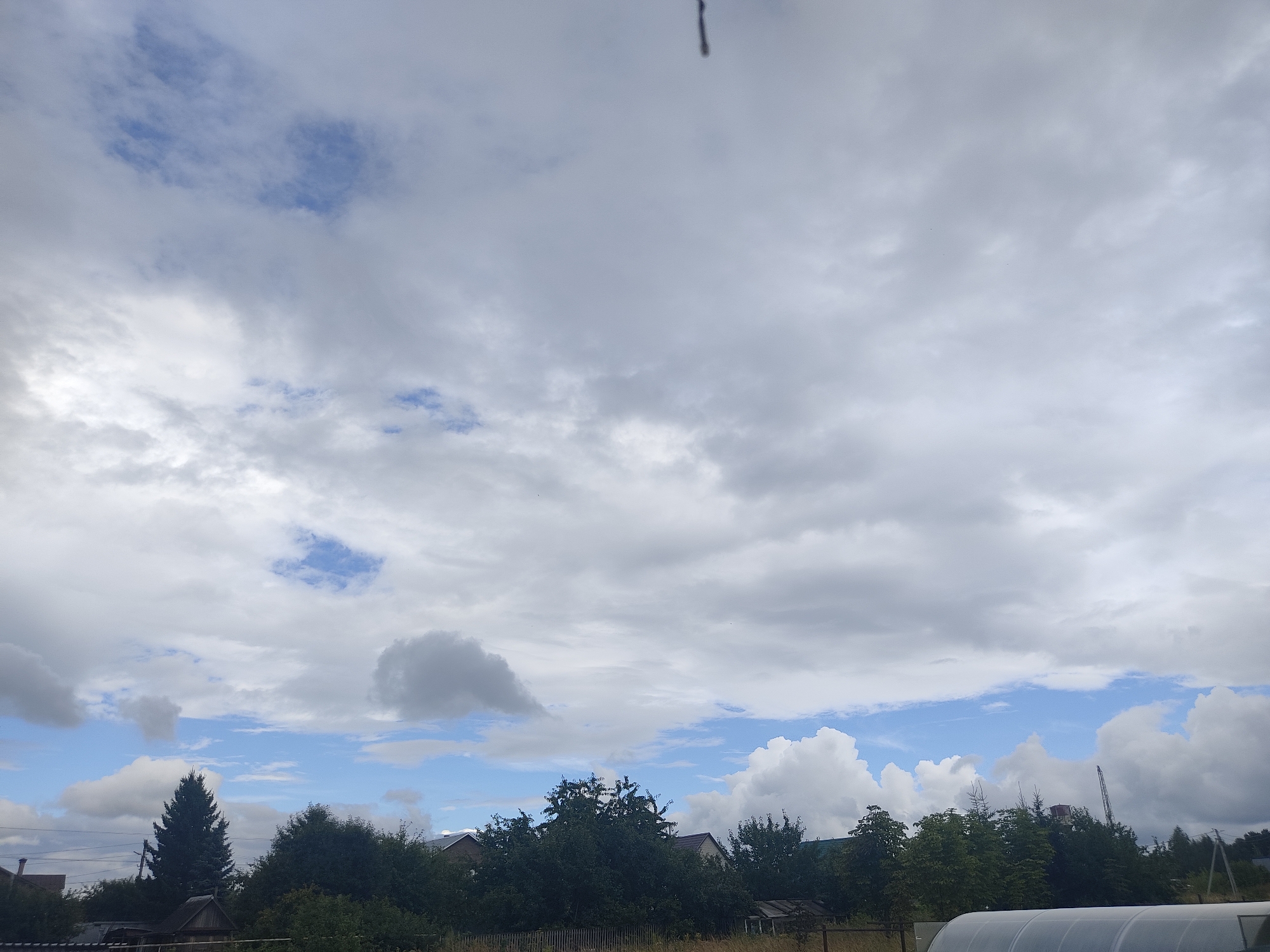 The image captures a cloudy sky over a rural area, possibly the village of Kamayka, Russia. The sky is a mix of white and gray clouds, with patches of blue sky visible. Below the sky, there is a dense line of trees, with some houses and structures peeking through. In the foreground, there are glimpses of a fence and a greenhouse-like structure. The overall impression is of a serene, overcast day in a countryside setting.