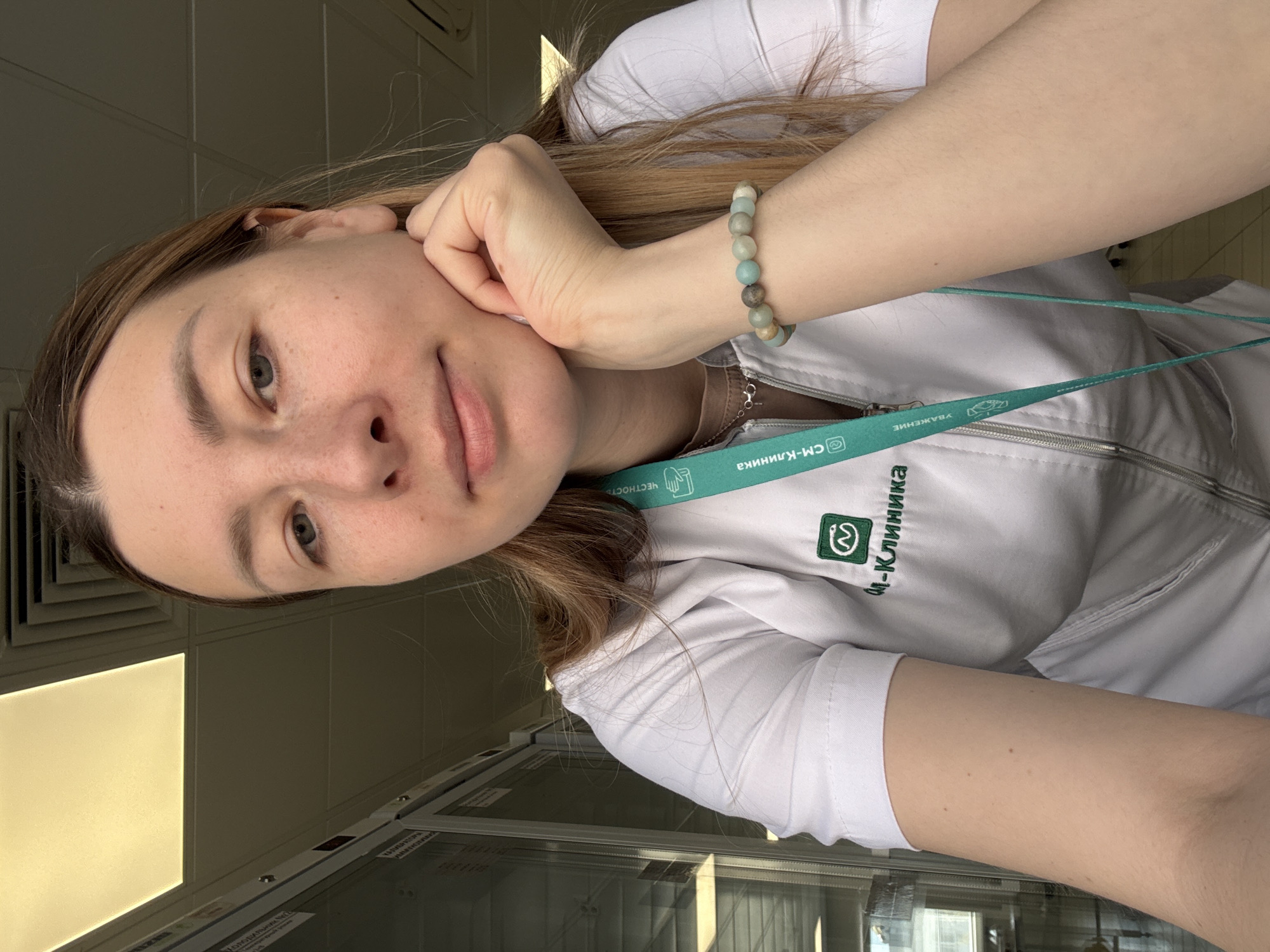 A woman is pictured indoors. She has light brown hair and gray-green eyes. She is wearing a white medical coat with a green lanyard, which reads "CM-Клиника" in Cyrillic letters, which is a medical clinic chain in Moscow, Russia. Her right hand is near her face. She is wearing a beaded bracelet on her right wrist. The background includes a window with a gray tile wall, indicative of a clinical or office environment.