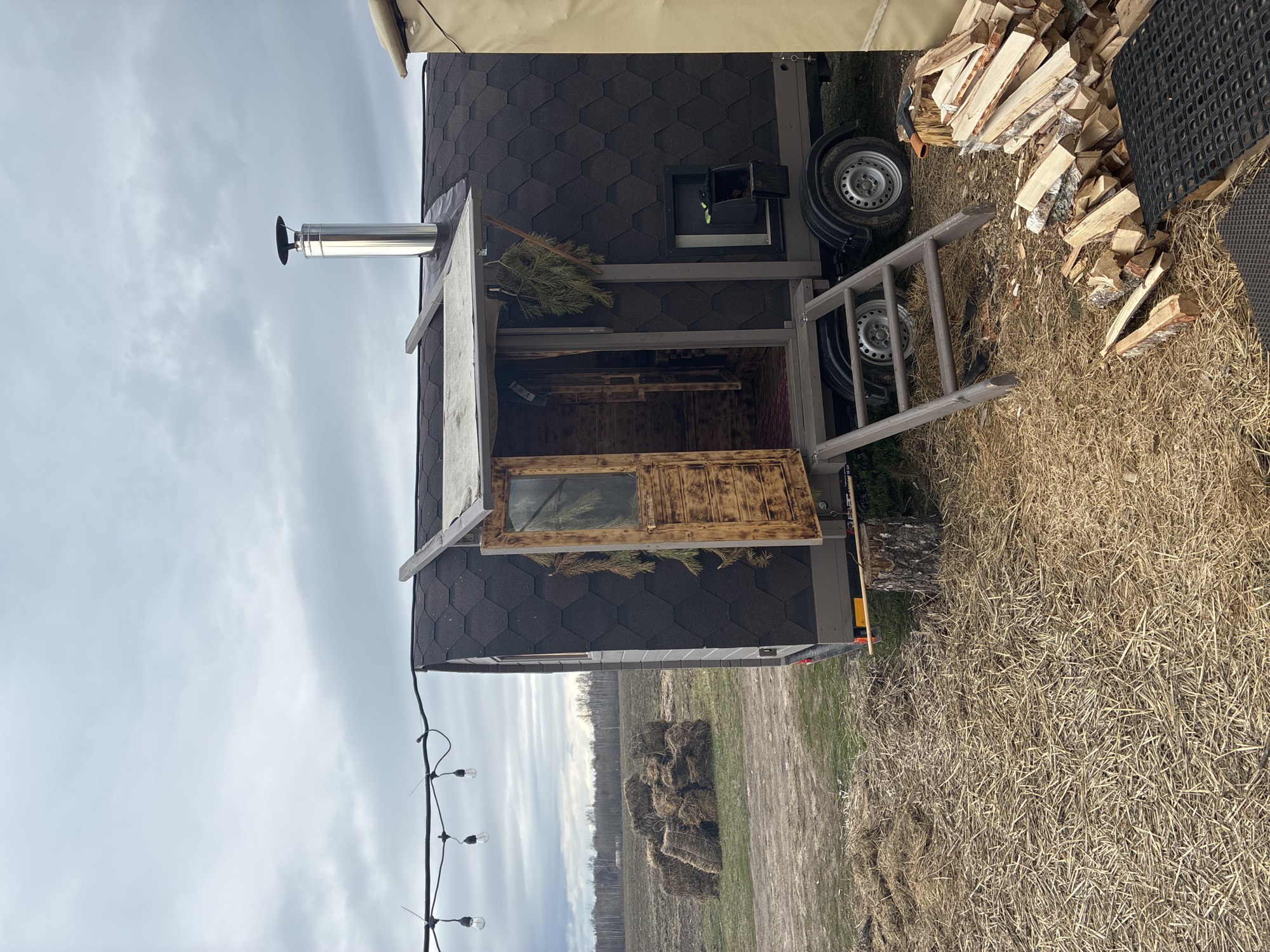 The image depicts a mobile sauna located in an outdoor environment, likely a rural or semi-rural area, evidenced by the presence of dry grass, hay bales in the background, and a wooded area in the distance. The sauna is constructed with dark-colored roofing shingles and light gray siding. A metal chimney protrudes from the top of the sauna. A wooden door is open, revealing the interior. A ladder rests against the side of the sauna, near two wheels of the trailer on which the sauna is mounted. A pile of split firewood is visible near the sauna. A string of light bulbs is visible on the lower left side of the image. The sky is overcast. The location is Unknown, Unknown.