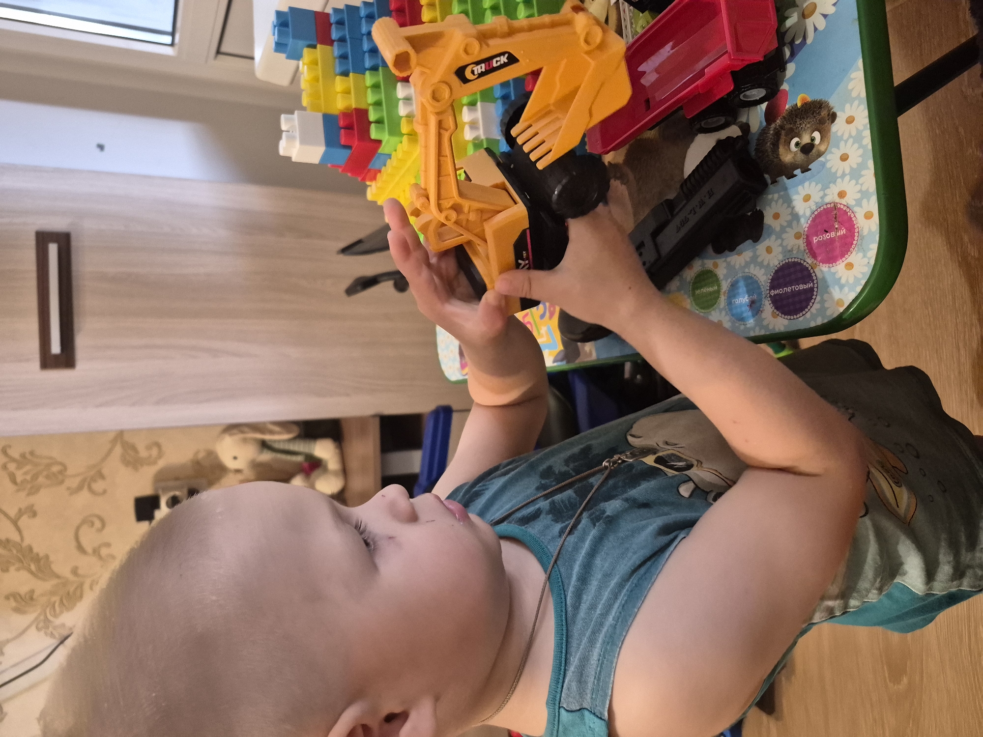 The image shows a close-up of a young child's face and hands interacting with toys on a table. The child, who appears to be a toddler, is wearing a teal tank top with a graphic print. They are holding a yellow toy excavator with black wheels. In the background, a collection of colorful building blocks, a red toy truck, and a small stuffed hedgehog are visible on the table. The table surface has a colorful pattern of daisies. A part of a window frame and a wooden floor are also visible.
The child's expression is focused on the toy, with their tongue slightly sticking out, suggesting concentration or engagement. The scene appears to be indoors, possibly a playroom or living area. The lighting suggests it could be daytime.
The visible text on the yellow toy excavator reads "TRUCK". There are also circular stickers on the table with Russian text, including "розовый" (pink), "зеленый" (green), "голубой" (light blue), and "фиолетовый" (purple).