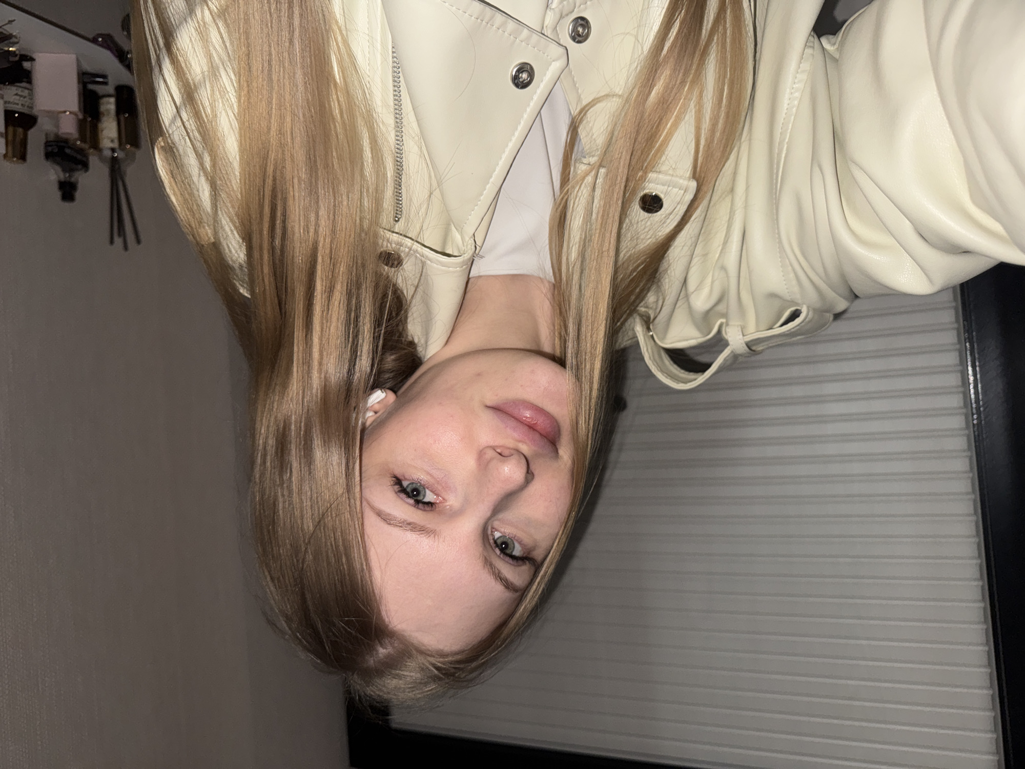 A young woman is captured in a selfie-style portrait, looking directly at the camera with a neutral to slightly smiling expression. She has long, straight, light brown hair that frames her face, fair skin, and light-colored eyes. She is wearing a light-colored, possibly cream or off-white, leather-like jacket with prominent snap buttons and a visible zipper detail, over a plain white top. A white earbud is visible in her left ear.
The setting appears to be indoors, likely a room in a home. The background to her left features a plain, light grey wall. To her right, a horizontally striped surface is visible, which could be window blinds or a radiator cover, also in a light grey tone. In the upper left portion of the image, several bottles, possibly cosmetics or perfumes, are arranged on a shelf or surface, alongside a reed diffuser. The lighting suggests an indoor, possibly evening or low-light environment, with artificial illumination. No text is visible in the image.