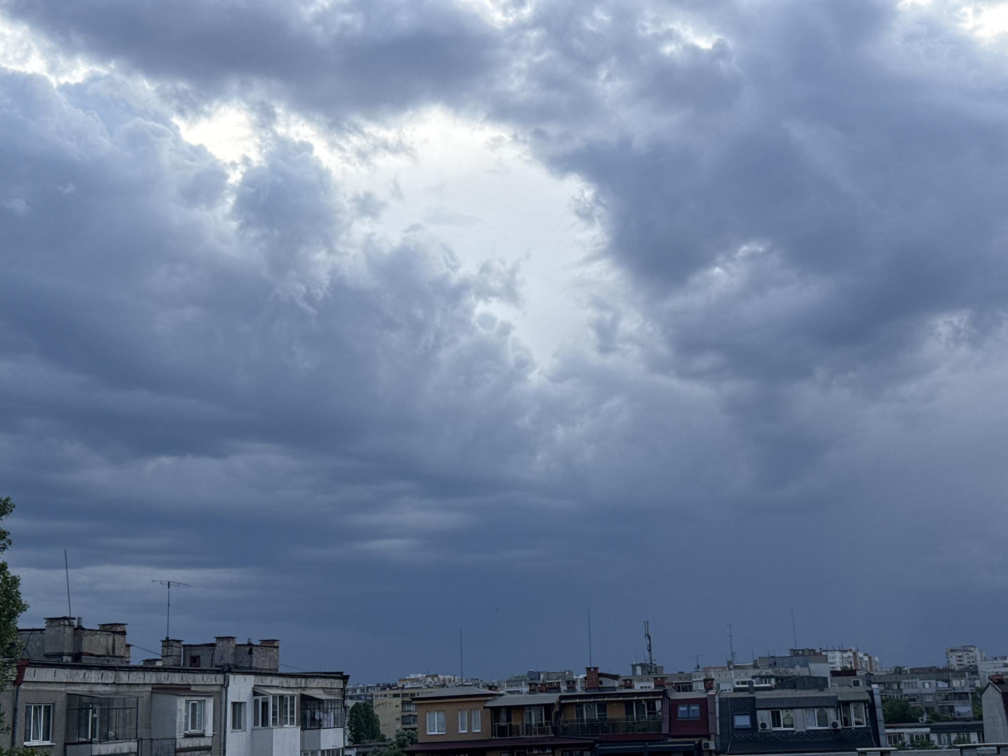 The image captures an urban or suburban landscape under a dramatically overcast sky. The upper two-thirds of the frame are dominated by heavy, dark grey storm clouds, suggesting impending rain or recent stormy weather. A distinct, irregularly shaped opening in the center of the sky reveals a brighter, lighter grey or white patch, where the clouds are thinner, hinting at a break in the cloud cover or a distant light source.
Below the expansive sky, the lower third of the image features a dense collection of buildings, primarily residential or mixed-use structures, stretching across the horizon. These buildings are multi-story, with flat roofs dotted with numerous chimneys, ventilation systems, and various antennae, including what appear to be television aerials and satellite dishes. The architecture suggests a utilitarian style, with some buildings featuring enclosed balconies or small extensions. Colors are generally muted, consisting of shades of grey, beige, and some darker red-brown roofs. A small section of green tree foliage is visible on the far left, partially obscuring one of the buildings.
The lighting is dim and diffused, characteristic of a heavily cloudy day. There are no people visible, and no discernible text. The scene evokes a sense of quiet urban life under a dramatic and unsettled sky.