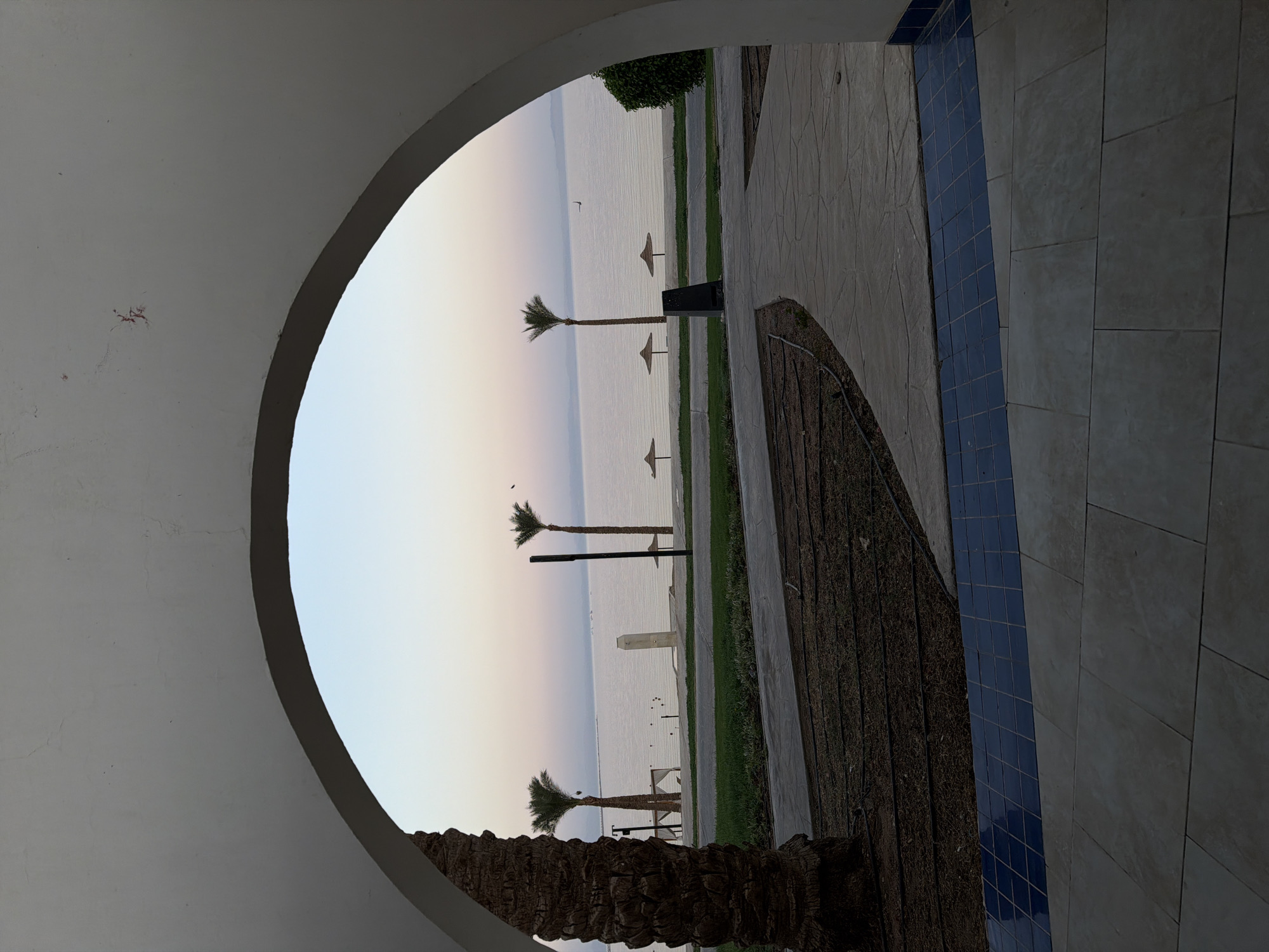 The image depicts a tranquil scene at dawn or dusk, looking through a white archway towards a body of water, likely the Red Sea, in Dahab, Egypt. The sky exhibits soft pastel hues of pink, purple, and pale blue. Several palm trees are visible, with one prominent one on the left side framing the view. On the shore, there are three straw-colored umbrellas, suggesting a beach or resort area. The water is calm, with a few small boats or objects visible in the distance. A modern outdoor shower fixture stands on the embankment. The foreground shows a paved walkway with a textured surface, bordered by dark soil with visible irrigation lines and a strip of blue tiles. The overall atmosphere is serene and suggests a peaceful vacation setting. There are no people visible in the frame.