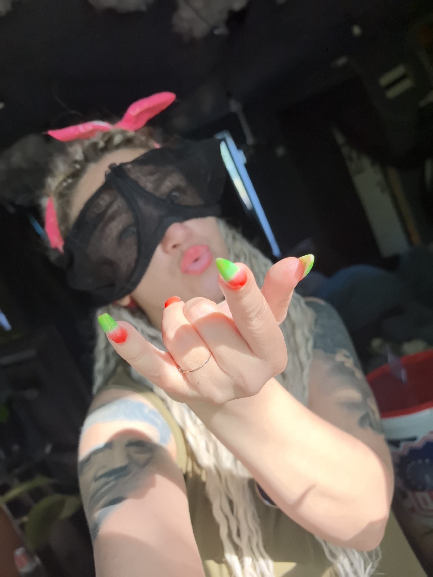 A person with long, curly blonde hair is indoors, wearing a pair of pink bunny ears and a black mesh bra over their eyes. They are making a hand gesture with their right hand, revealing a ring on their ring finger and nails painted in a green-to-red ombre style. Visible tattoos cover their arm. The background is dark and cluttered, with hints of furniture and an open window. The environment is indoor. The location is not determinable based on this image.