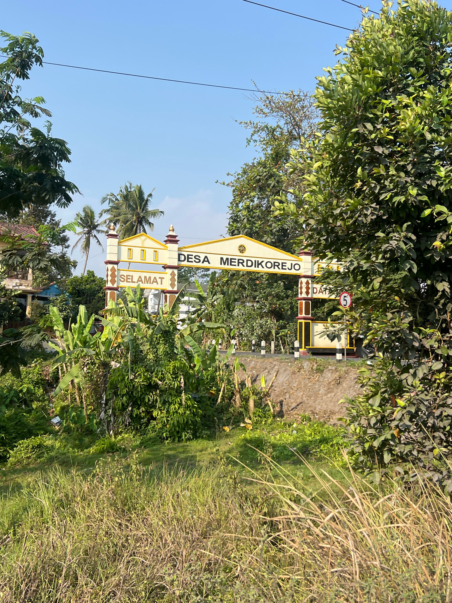 The image shows a welcome archway at the entrance to Desa Merdikorejo in Tempel, Indonesia. The archway is yellow and white with brown accents and features the words "SELAMAT" on the left and "DESA MERDIKOREJO" on the central banner. There is also text on the right pillar that reads "DAT" and a speed limit sign showing "5". The archway is surrounded by lush green vegetation, including banana trees and other tropical plants. In the background, there are palm trees and a clear blue sky. The ground appears to be a dirt road leading into the village. The scene is well-lit, suggesting it was taken during the daytime. There are no people visible in the image.