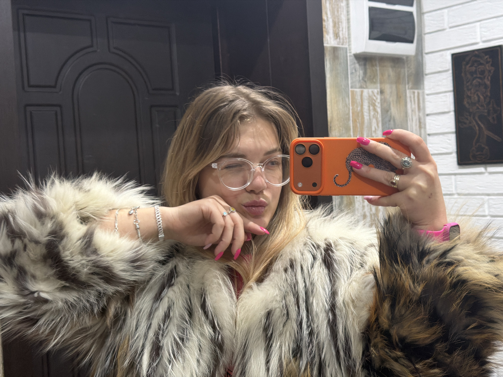 A young woman with light-colored hair and clear-framed glasses takes a mirror selfie indoors. She is seen from the chest up, wearing a thick, fluffy fur coat with varied shades of white, grey, brown, and black. Her fingernails are painted a vibrant pink, and she wears several silver rings and bracelets. She holds an orange smartphone, featuring a leopard print design and a triple camera array, to capture the image. The woman winks with her left eye and puckers her lips, conveying a playful expression.
The background on the left side of the image features a dark, paneled door with an arched design. To the right, the wall is composed of a white brick texture, adjacent to a section of patterned grey and brown tiling. On the white brick wall, a dark framed artwork depicting a bearded male bust is visible. The setting appears to be an interior space. No discernible text is visible within the image.