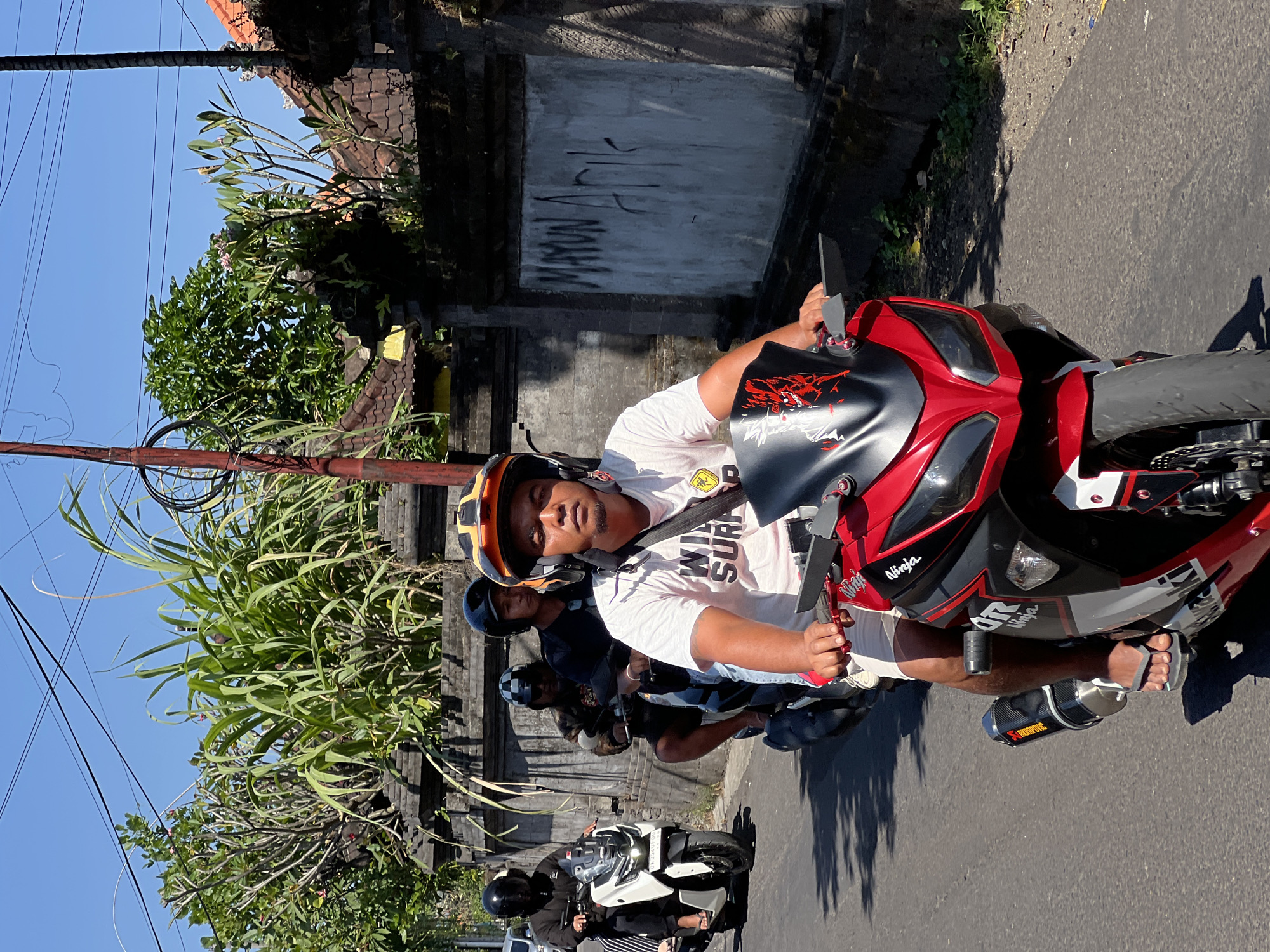 In Denpasar, Indonesia, on a sunny day, four men ride motorcycles in single file on a paved road. The man in the foreground, wearing a white t-shirt with "WILD SURFER" printed on it, an orange and yellow helmet, and white shorts, drives a red and black sports motorcycle. Behind him, a man in a black helmet rides a dark motorcycle. Further back, a man in a checkered helmet also rides a dark motorcycle, and in the distance, a man in a black helmet on a white scooter is visible. The road is bordered by a stone wall covered in vegetation and graffiti, with power lines overhead. The background features trees and buildings with tiled roofs, typical of the urban landscape in Denpasar. The overall scene depicts everyday street life in the city.