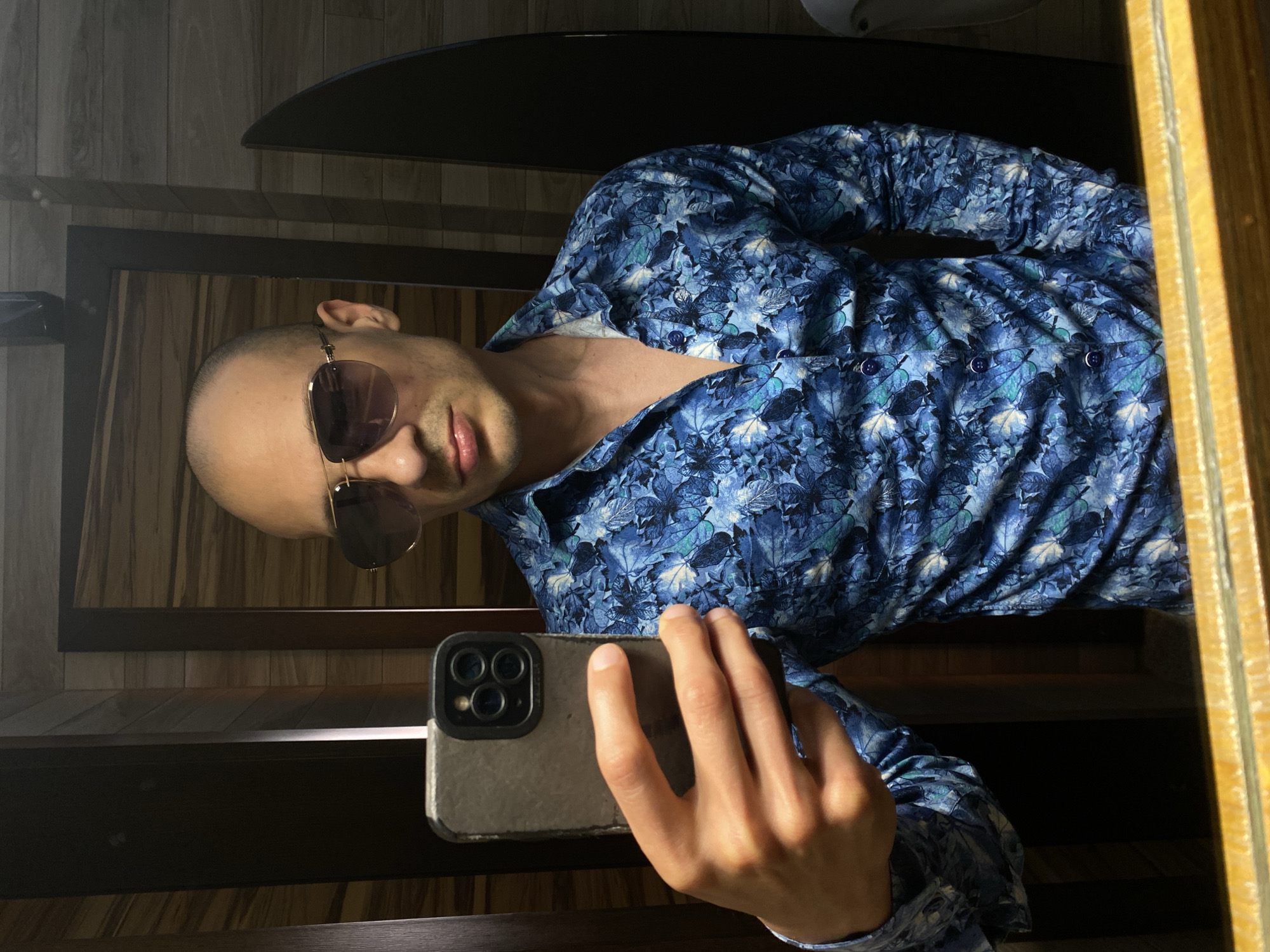 A person is holding a smartphone in their right hand, taking a selfie in front of a mirror. The person has a shaved head and is wearing sunglasses and a blue button-down shirt with a leaf pattern. The background appears to be a wooden interior, possibly a bathroom. The location is not discernible from the image and cannot be identified as Pleven, Bulgaria.