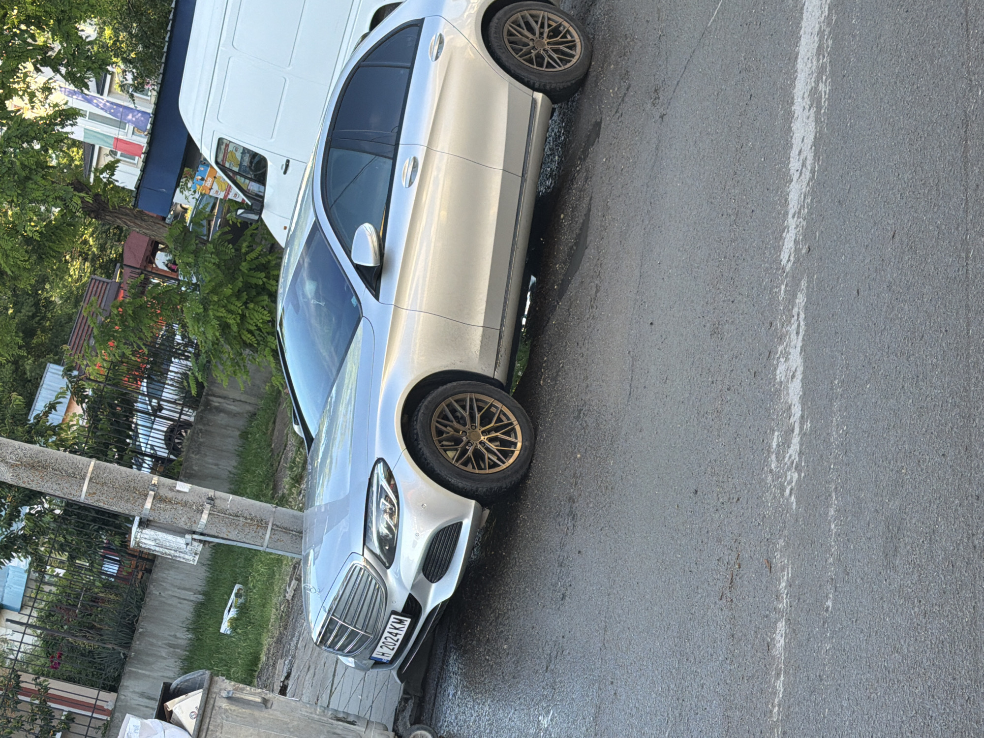 The image captures a silver Mercedes-Benz sedan parked on the side of a wet, asphalt road in what appears to be an urban or semi-urban setting during daytime. The car features distinctive bronze/gold multi-spoke alloy wheels. Its front license plate, "H 2024 KM," is clearly visible. The road surface shows patches of moisture or dark streaks.
To the left of the car, a concrete utility pole is positioned next to a paved area and a fence, with lush green foliage and trees in the background. A partially visible grey garbage bin is in the lower left corner.
Further down the street, a white commercial van is parked, partially obscured by trees and the main car. A "BILLA" logo is visible on the side of the van, suggesting it may belong to the supermarket chain. In the background, on a building, flags are visible, including what appears to be the Bulgarian national flag and the European Union flag, providing a clear location cue for Shumen, Bulgaria. No people are visible, and the scene depicts parked vehicles on a bright, clear day, despite the damp road.