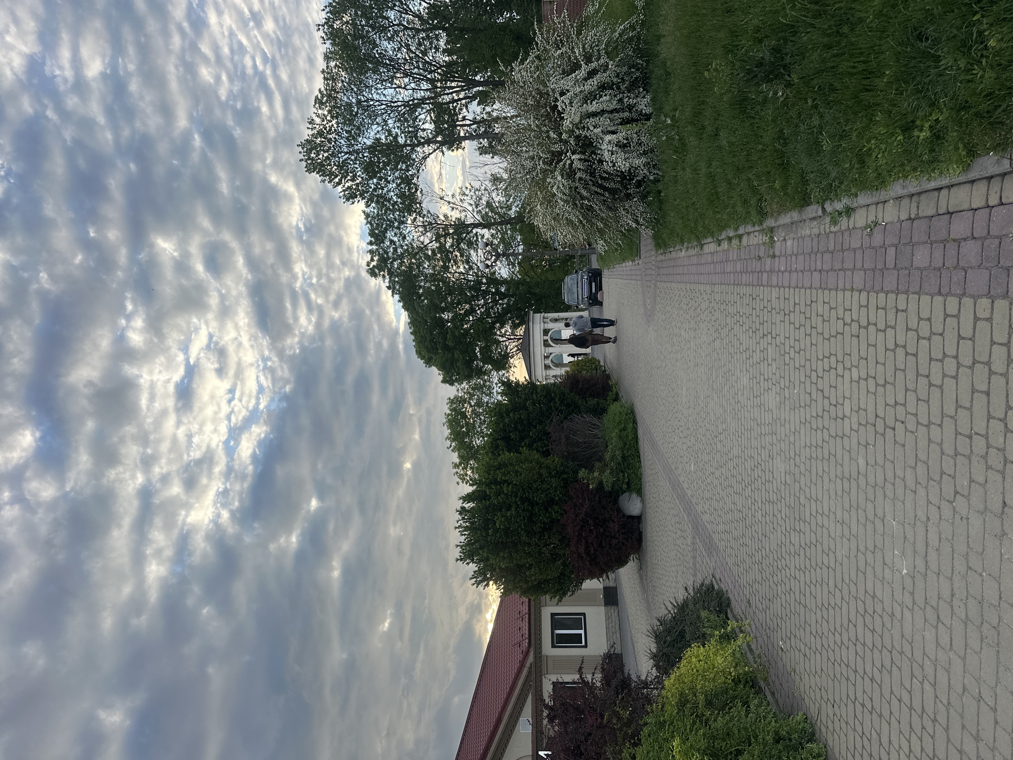 The image depicts an outdoor scene in Ternopil, Ukraine. The primary focus is a paved walkway constructed of interlocking pavers, flanked by green grass and landscaping.
A building with a red tile roof and white walls is visible on the lower right of the image. Several people are walking along the path in the distance, near a white structure with columns and a parked black SUV.
The sky is dominated by cloudy, textured formations, suggesting an overcast day or nearing sunset. The light filtering through the clouds creates an atmospheric effect.