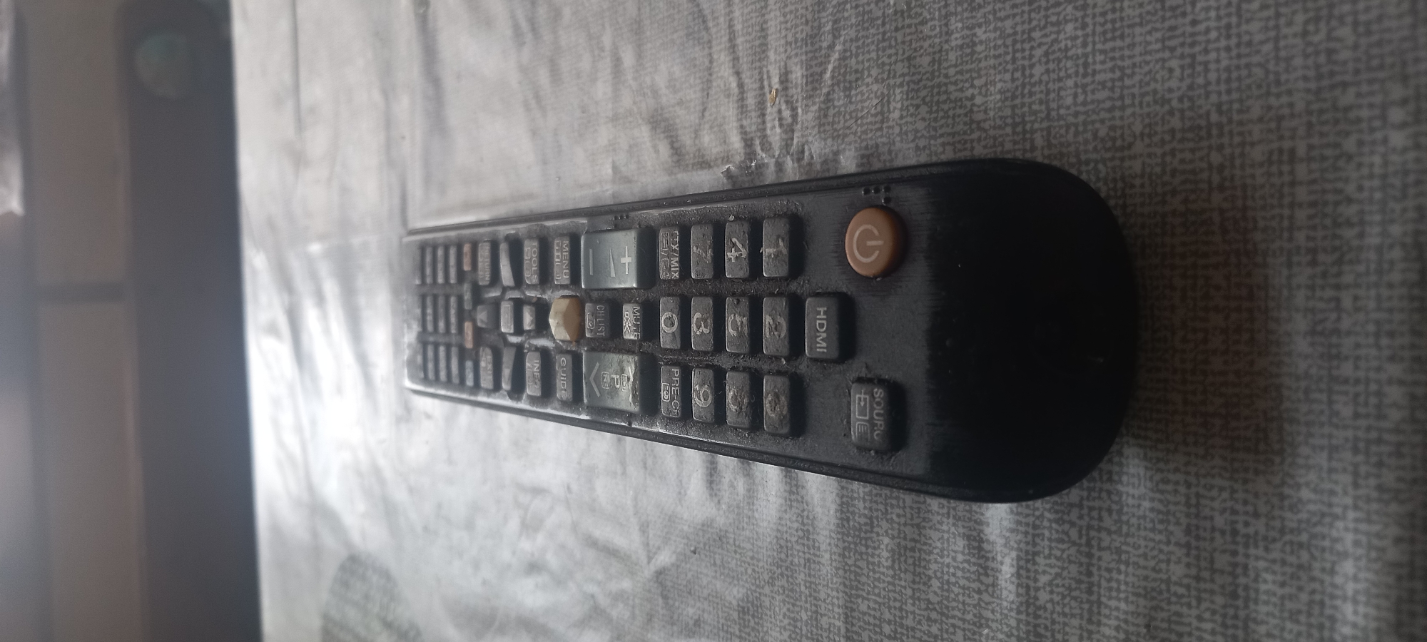 The image shows a black Samsung TV remote lying on a gray patterned surface. The remote is dusty and shows signs of wear. The buttons are visible, including power, HDMI, volume, channel, menu, and number buttons. The overall composition suggests a domestic setting. The location of the image is unknown.