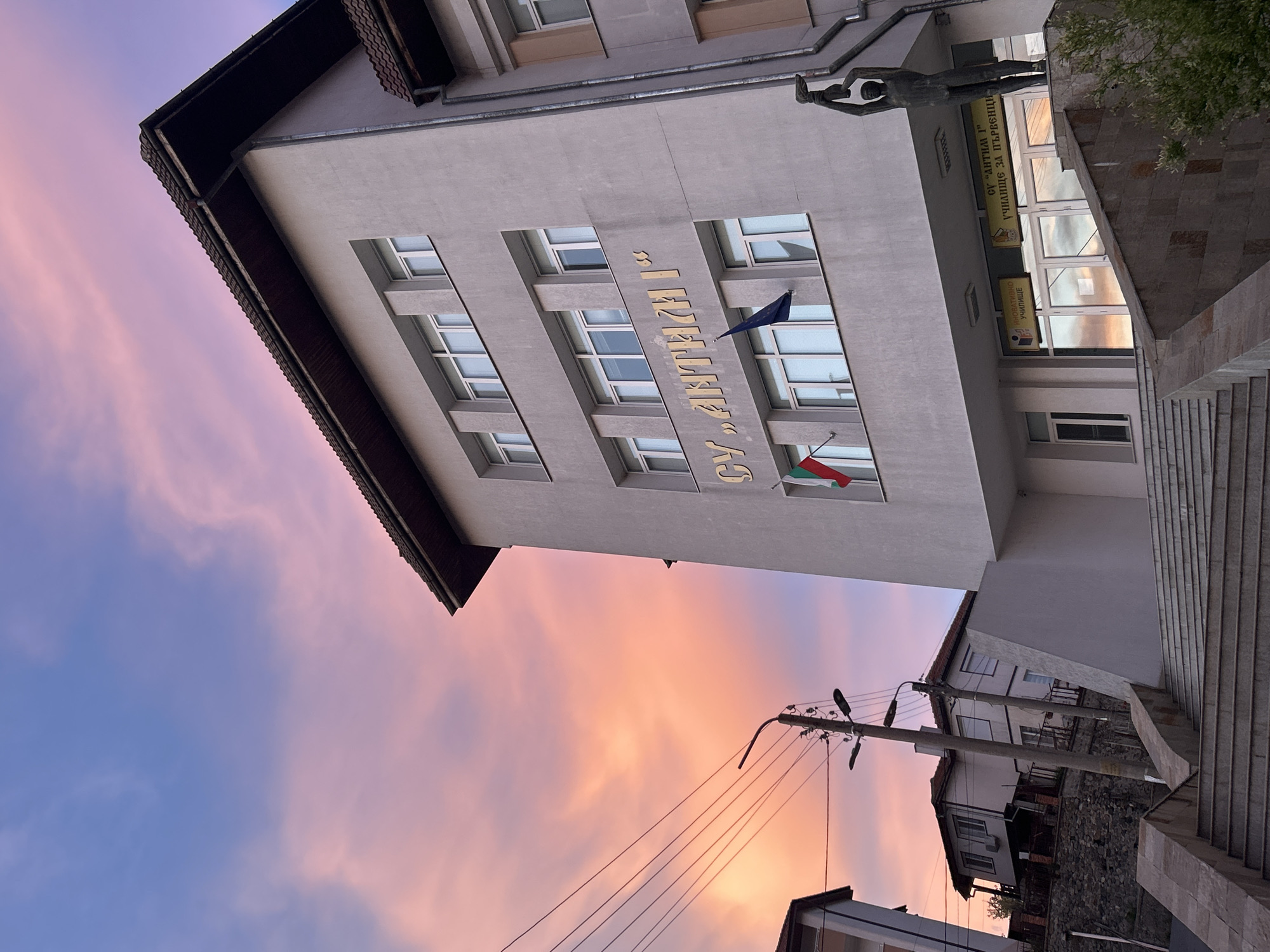 This photo, taken in Zlatograd, Bulgaria, shows a low-angle, off-center view of a building under a sky with pink and blue clouds. The building has a light gray facade with a multi-paned window and is topped with a brown tiled roof. The words "СУ „АНТИМ I“" are inscribed in gold letters. The flag of Bulgaria and the European Union are attached to the building. To the right of the building, a stone staircase ascends. A sign is to the right of the staircase, with the words "ОУ "АНТИМ I" УЧИЛИЩЕ ЗА ПЪРВЕНЦИ" which translates to "OU "ANTIM I" School for Firsts". Above the sign is a bronze statue of two figures. In the lower part of the photograph, there are other buildings and a utility pole with wires. The lighting indicates either sunrise or sunset.