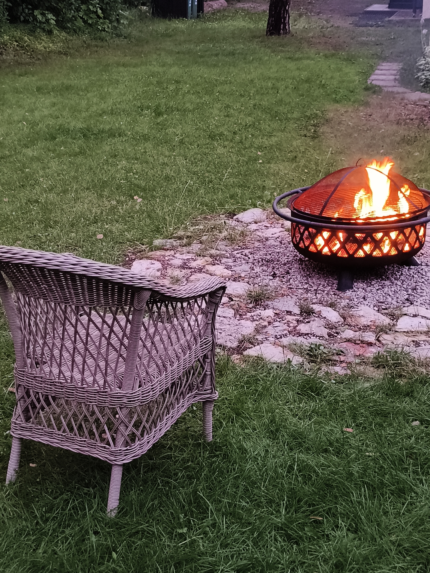 In Bigauņciems, Latvia, a fire pit with active flames is situated on a gravel and stone patio. Adjacent to the patio, a wicker chair sits on a well-maintained lawn. The surrounding area is characterized by lush green grass and scattered foliage, suggesting an outdoor setting, possibly a backyard or garden. The scene appears to be set in the evening, as indicated by the ambient light and the glowing embers within the fire pit.