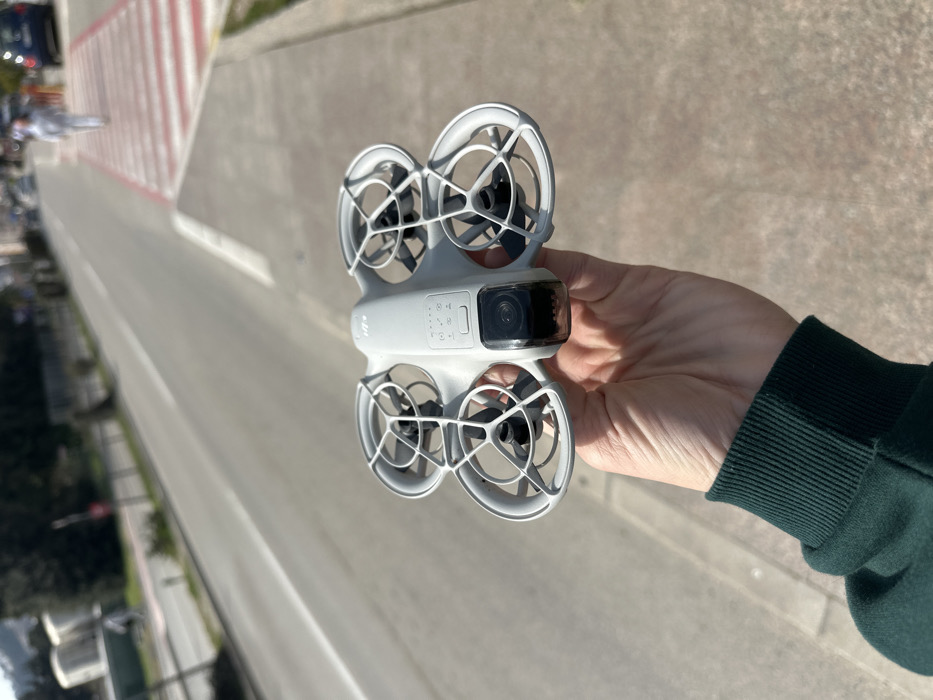 A first-person view image depicts a light gray DJI Avata-style FPV drone, equipped with integrated propeller guards and a forward-facing camera, held horizontally in an open right hand. The hand is positioned centrally in the lower frame, wearing a dark green long-sleeved garment.
The background presents an outdoor urban street scene under bright, direct sunlight. A two-lane asphalt road extends from the lower left foreground towards the upper right background, featuring a continuous white lane divider. Parallel to the road on the right, a paved sidewalk is visible. A distinctive pedestrian crosswalk with alternating red and white stripes is located further down the street, intersecting the road and sidewalk on the right.
A person dressed in light-colored clothing is observed walking away from the camera across this red and white striped crosswalk in the mid-distance. Blurred vehicles are faintly visible on the road in the far distance. The background also includes indistinct urban structures and distant, tree-covered hills under a clear blue sky.
The location is identified as a section of **Jadranski put (Adriatic Highway)** in Budva, Montenegro, specifically in the central urban area, likely near Slovenska Plaža or TQ Plaza, characterized by the unique red and white striped pedestrian crossing.