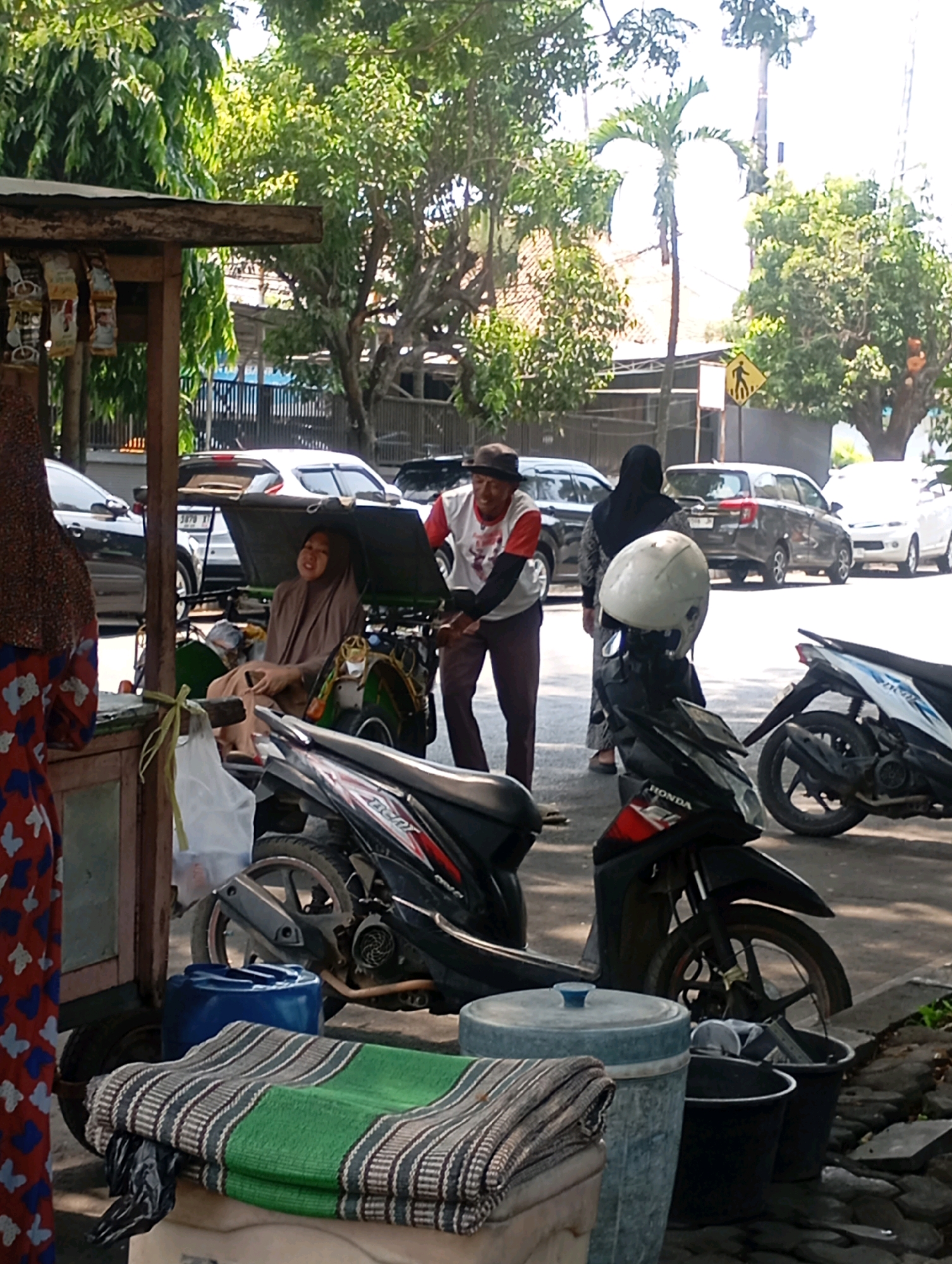 The image depicts a street scene in Sumber, Indonesia, with a woman wearing a hijab seated in a three-wheeled vehicle, possibly a bemo or a modified motorcycle. She is looking towards the camera. Beside her, a man in a red and white shirt and a hat stands, seemingly interacting with her. In the foreground, a woman with a colorful patterned dress is partially visible, standing near a wooden stall displaying various packaged goods. Several motorcycles are parked in the vicinity, including a black Honda. The background shows a street with cars, palm trees, and other vegetation, indicating an outdoor, possibly urban or semi-urban, environment. The lighting suggests it is daytime.