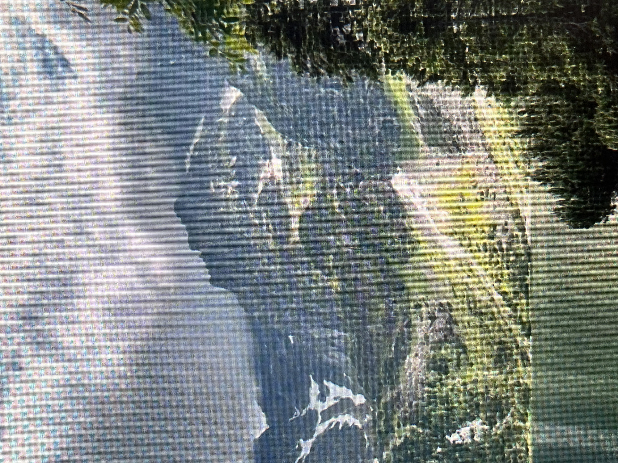 The image shows a landscape of mountains and a body of water. The mountains are rocky with some vegetation and snow patches. Clouds obscure parts of the mountain range. Trees are visible in the foreground, framing the scene. The location is not identifiable as Tbilisi, Georgia from the image alone.