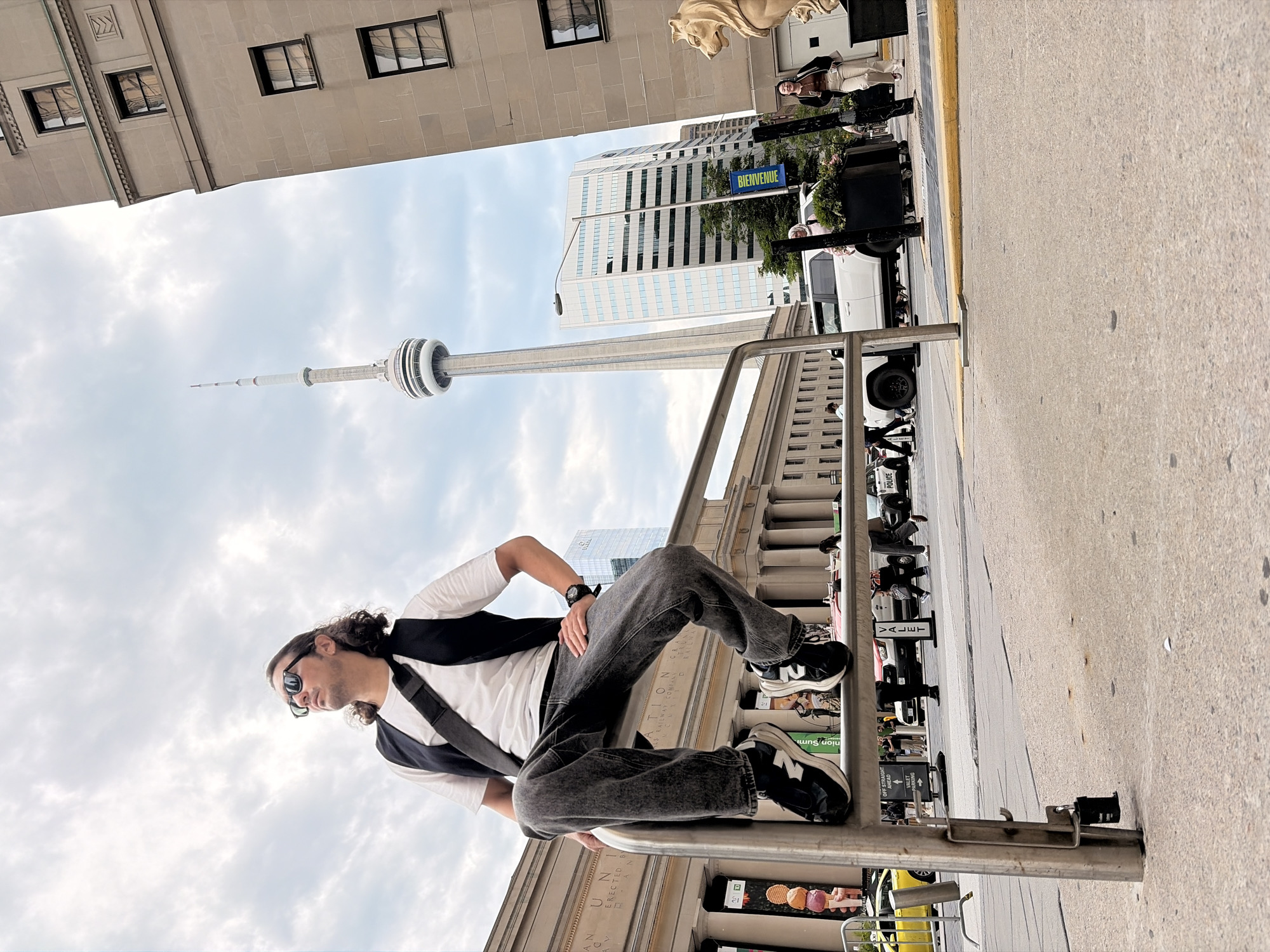 A man with long, curly brown hair and sunglasses is seated on a metal railing in front of a large building. He is wearing a white t-shirt, a dark vest, and dark grey jeans. He has a black crossbody bag slung over his shoulder. The man is looking up and to his left. In the background, the CN Tower is visible, along with other buildings and a street with traffic and pedestrians. The environment appears to be an urban setting in Toronto, Canada, likely near Union Station, given the architectural style of the building behind the man which resembles the historic train station. The sky is overcast with clouds.