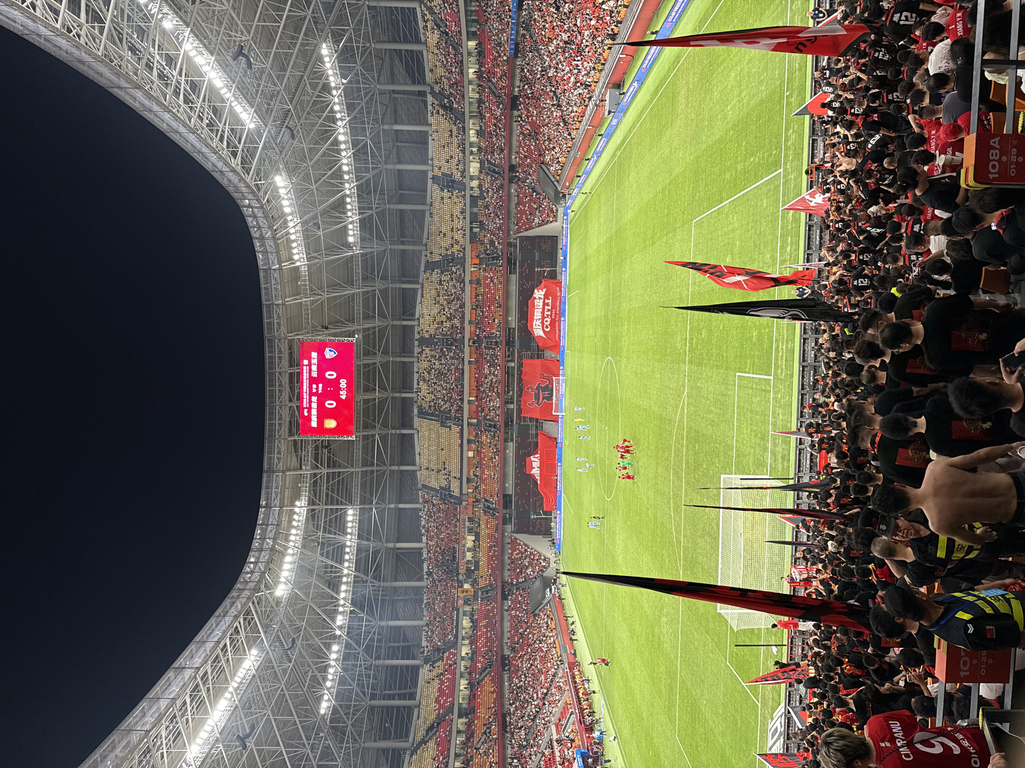 The image shows a soccer stadium with a game in progress. The stadium is full of spectators, with a large contingent of fans near the field waving red and black flags. A scoreboard indicates the game is at halftime, with the score at 0-0. A game between Chongqing Tonglianglong and Yunnan Yukun is indicated by text on a large banner. The stadium's roof structure is visible, as are the floodlights illuminating the field. The scene is presumably taken at Longxing, China, based on the team's location.